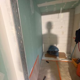 Szo Ne Bud - Wnętrze w trakcie prac wykończeniowych: zielone panele ścienne z widocznymi profilami montażowymi, częściowo wykończone ściany i sufit. Podłoga zabezpieczona, na ścianie cień osoby.