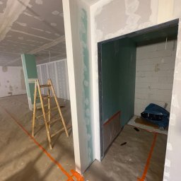 Szo Ne Bud - Wnętrze w trakcie prac wykończeniowych: ściany i sufit z płyt gipsowych, częściowo pokryte masą. Na podłodze pomarańczowe linie, w tle drewniana drabina i otwór drzwiowy.