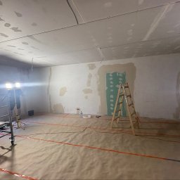 Szo Ne Bud - Remontowane wnętrze z sufitem podwieszanym i częściowo wygładzonymi ścianami. Na podłodze papier ochronny z pomarańczowymi liniami, rusztowanie, drabina i lampa robocza.
