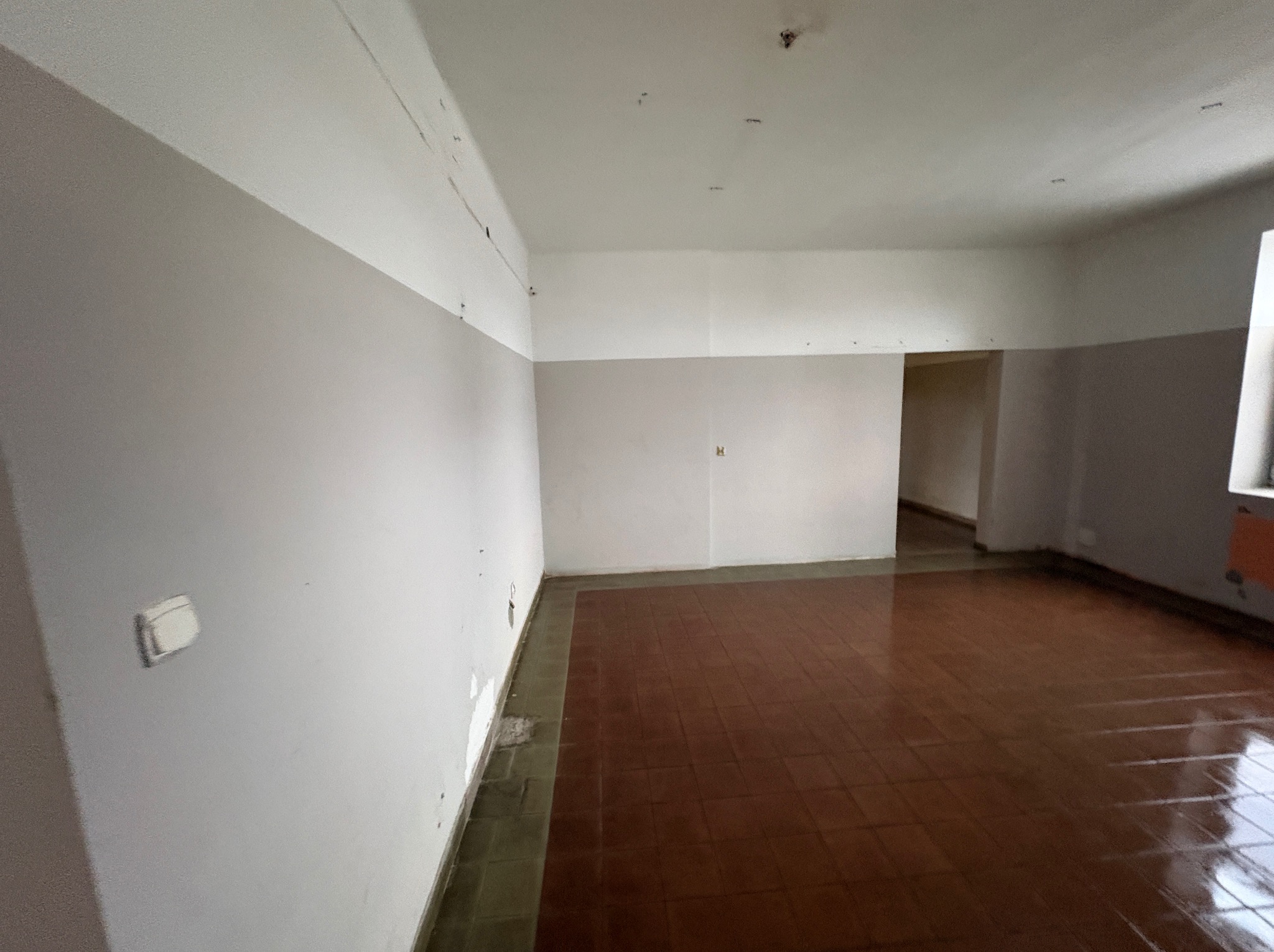 Pusty pokój przed remontem: szare ściany z widocznymi śladami, brązowa podłoga z płytek, biały sufit z wystającymi elementami. Widok na sąsiednie pomieszczenie.