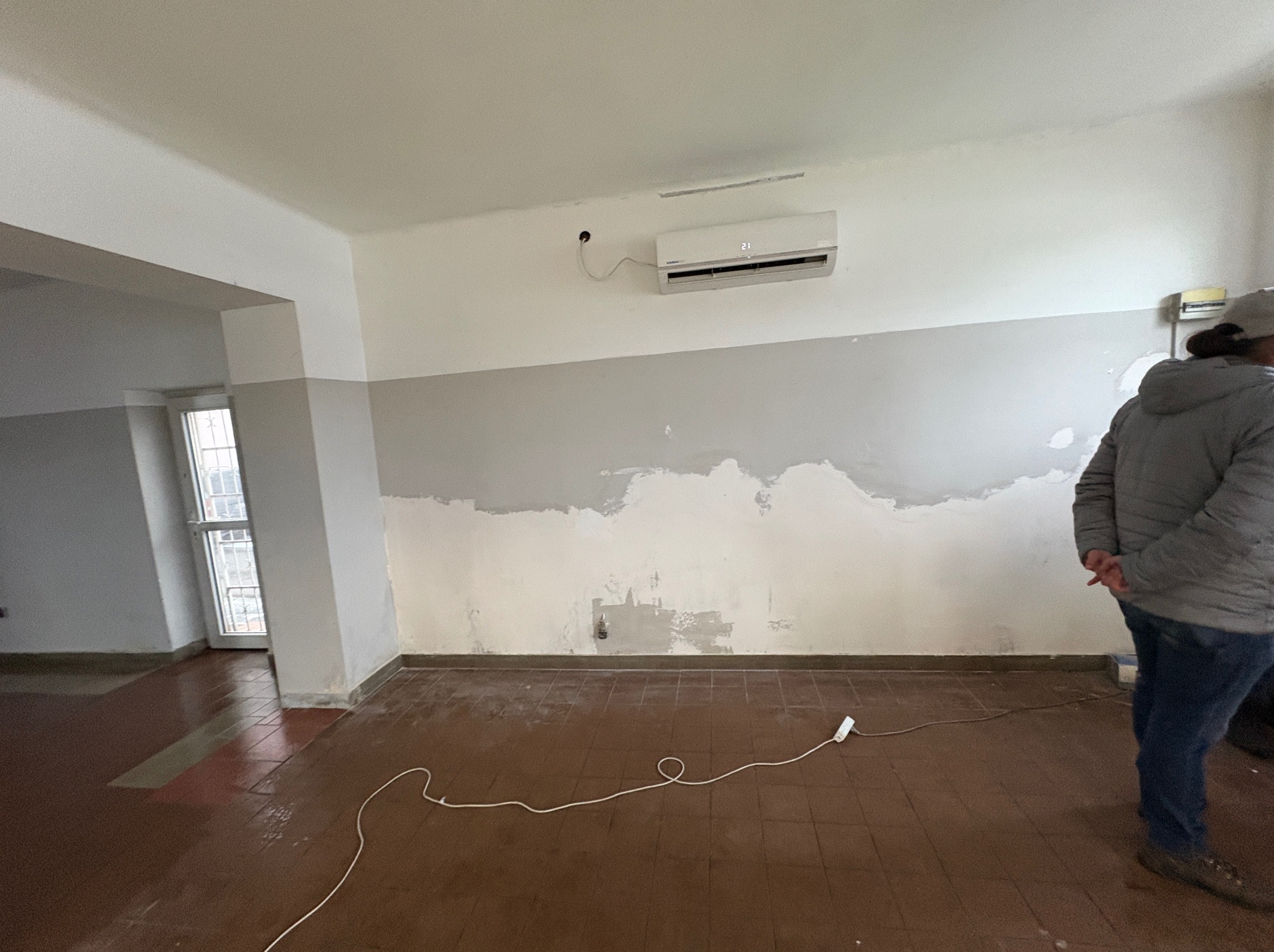 Pokój w trakcie szpachlowania: ściana z widocznymi łatami szpachli, klimatyzator na górze, brązowa podłoga z terakoty, kabel na podłodze, osoba w tle.