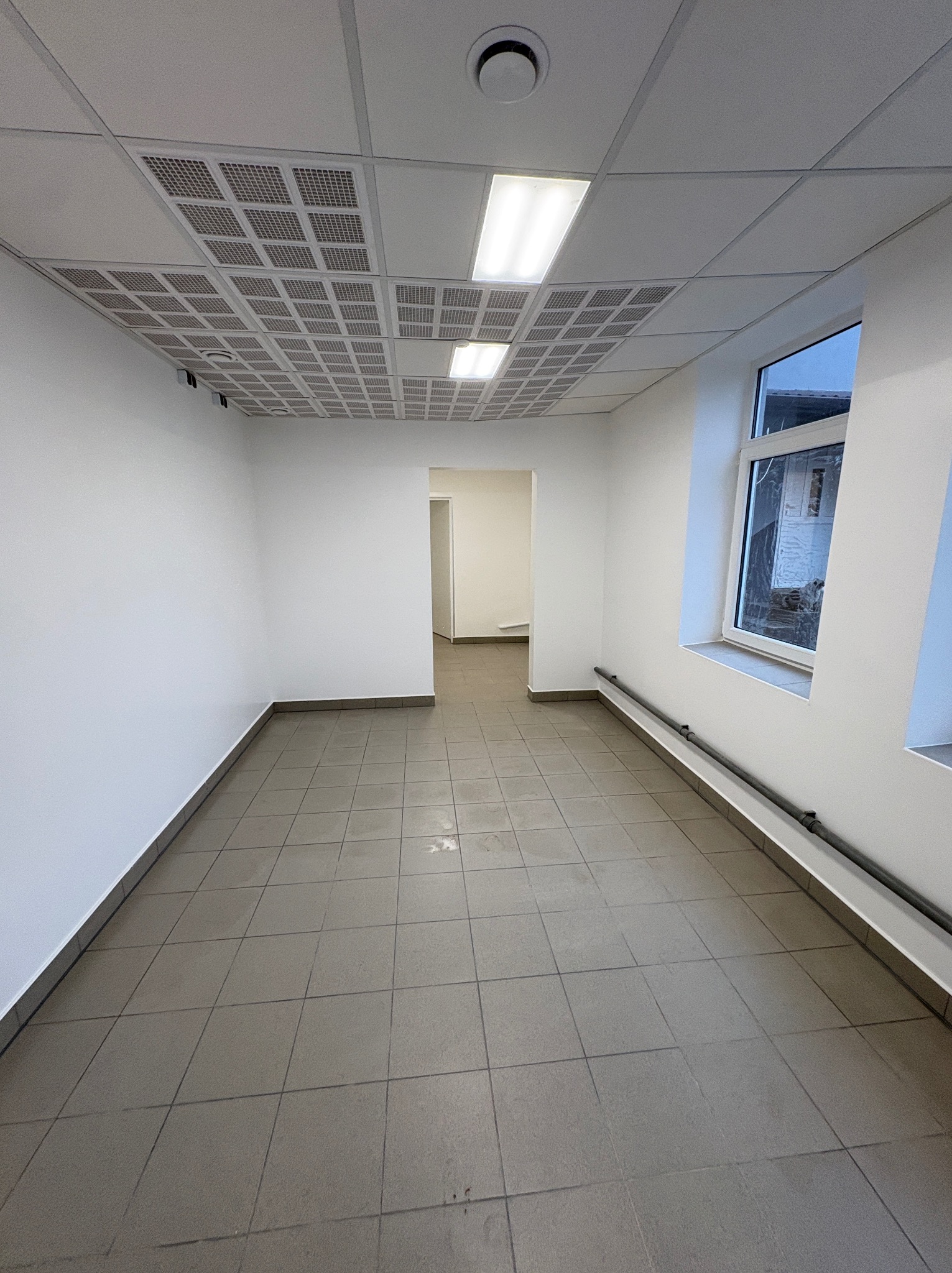 Wnętrze biura po remoncie w Warszawie: białe ściany, szara podłoga z płytek, sufit kasetonowy z oświetleniem. Widok na korytarz i okno.