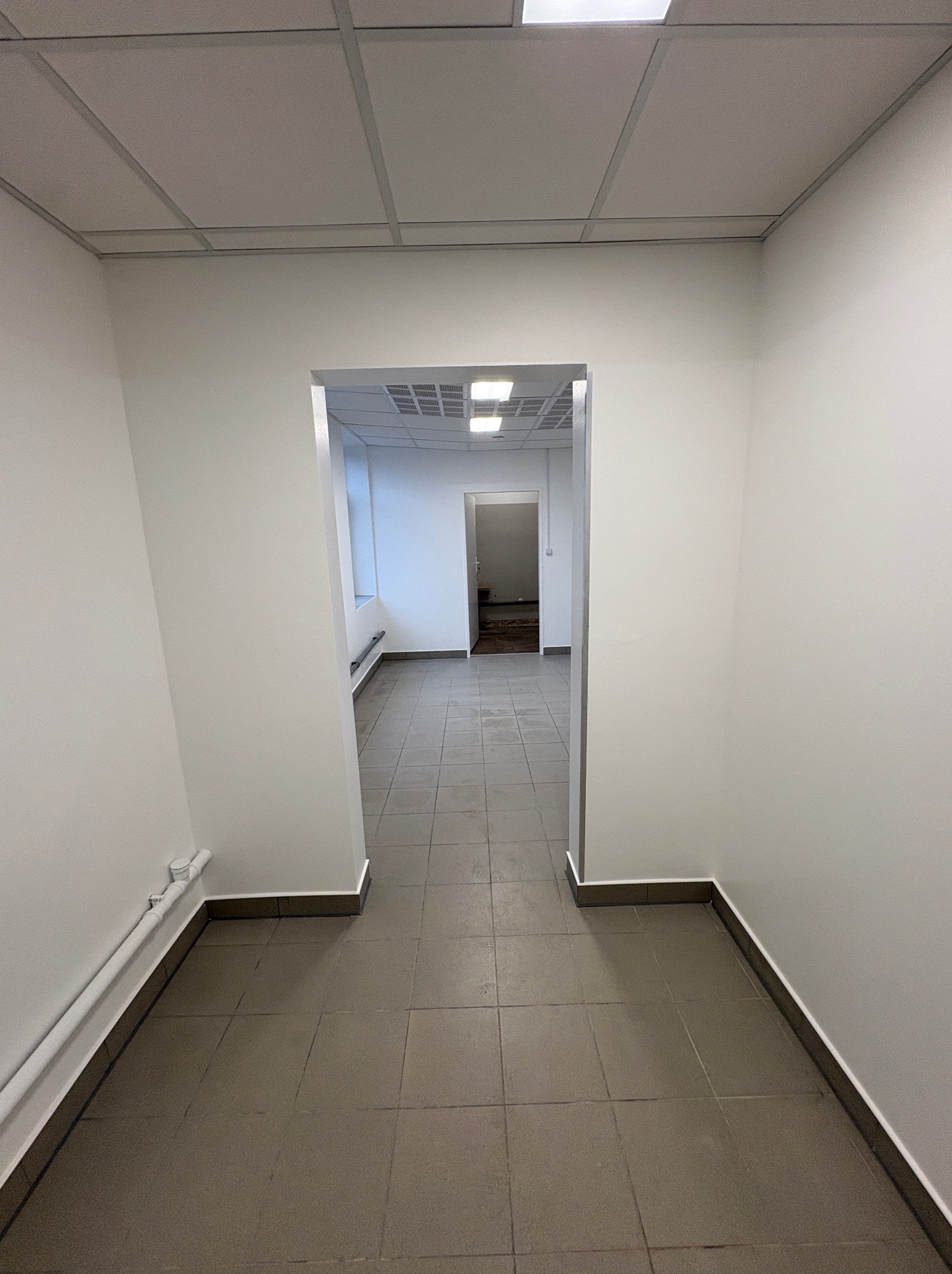 Korytarz biurowy z szarą podłogą i białymi ścianami, widok w głąb pomieszczenia przez otwarte drzwi. Sufit kasetonowy. Listwy przypodłogowe.