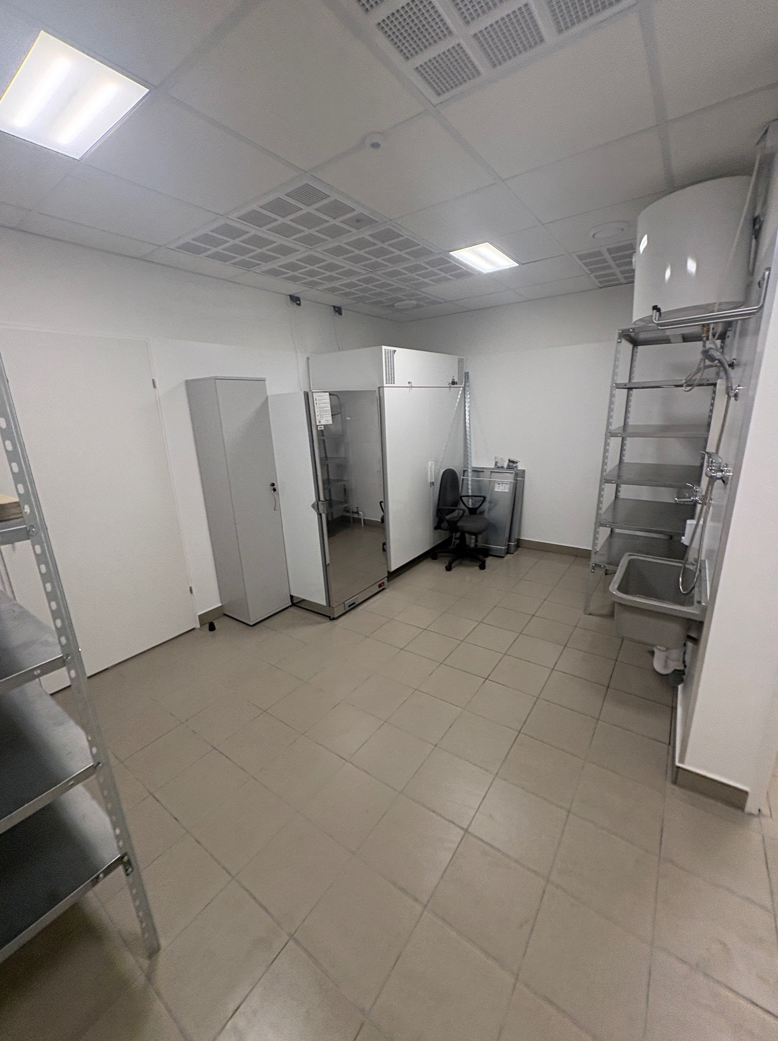 Białe pomieszczenie z płytkami podłogowymi, szafkami, umywalką i metalowymi regałami. Widok z perspektywy, oświetlenie sufitowe.