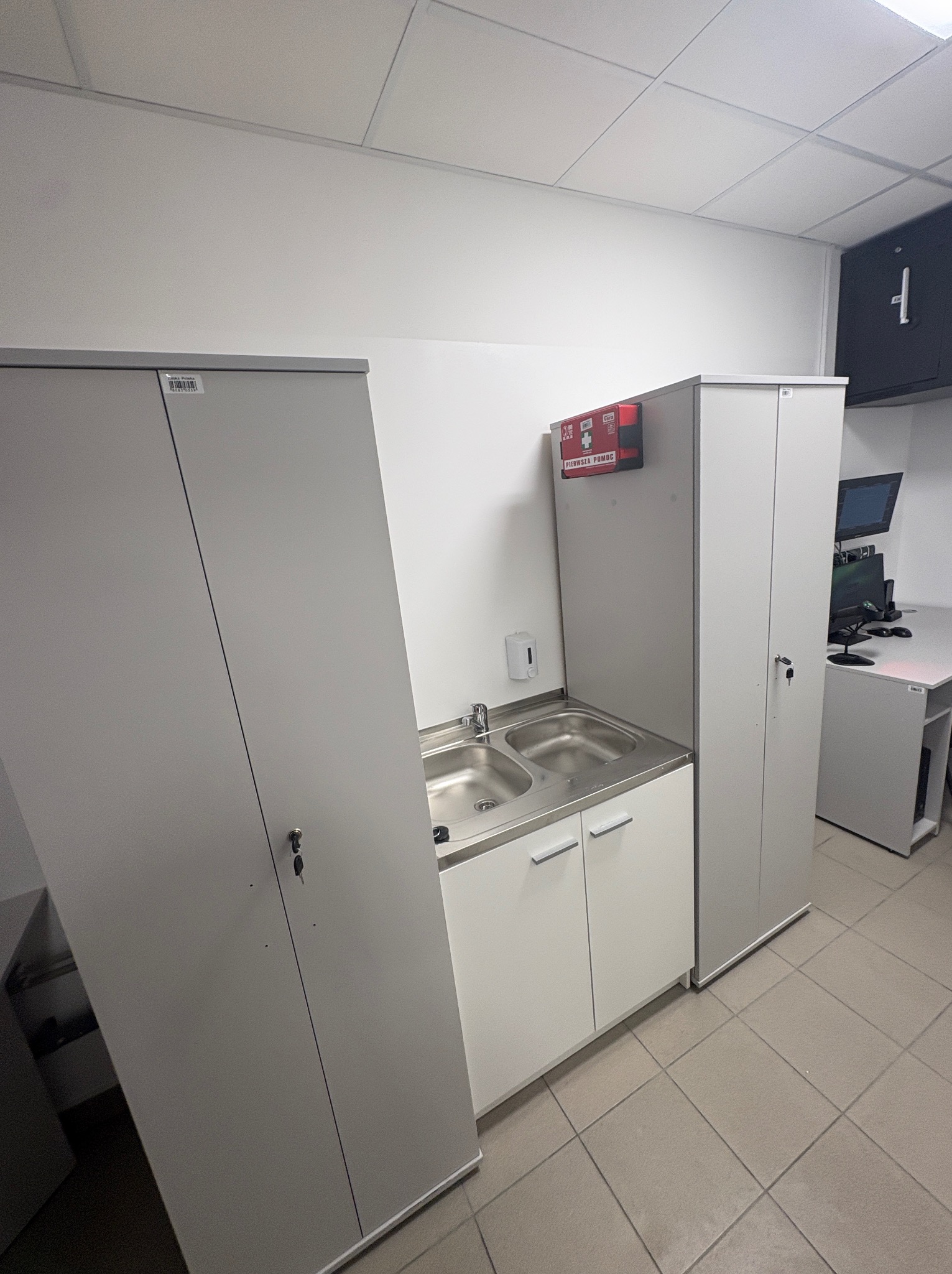 Wyposażenie socjalne biura po remoncie: szafy metalowe, umywalka z szafką, apteczka pierwszej pomocy. Widok na stanowiska pracy z komputerami w tle.