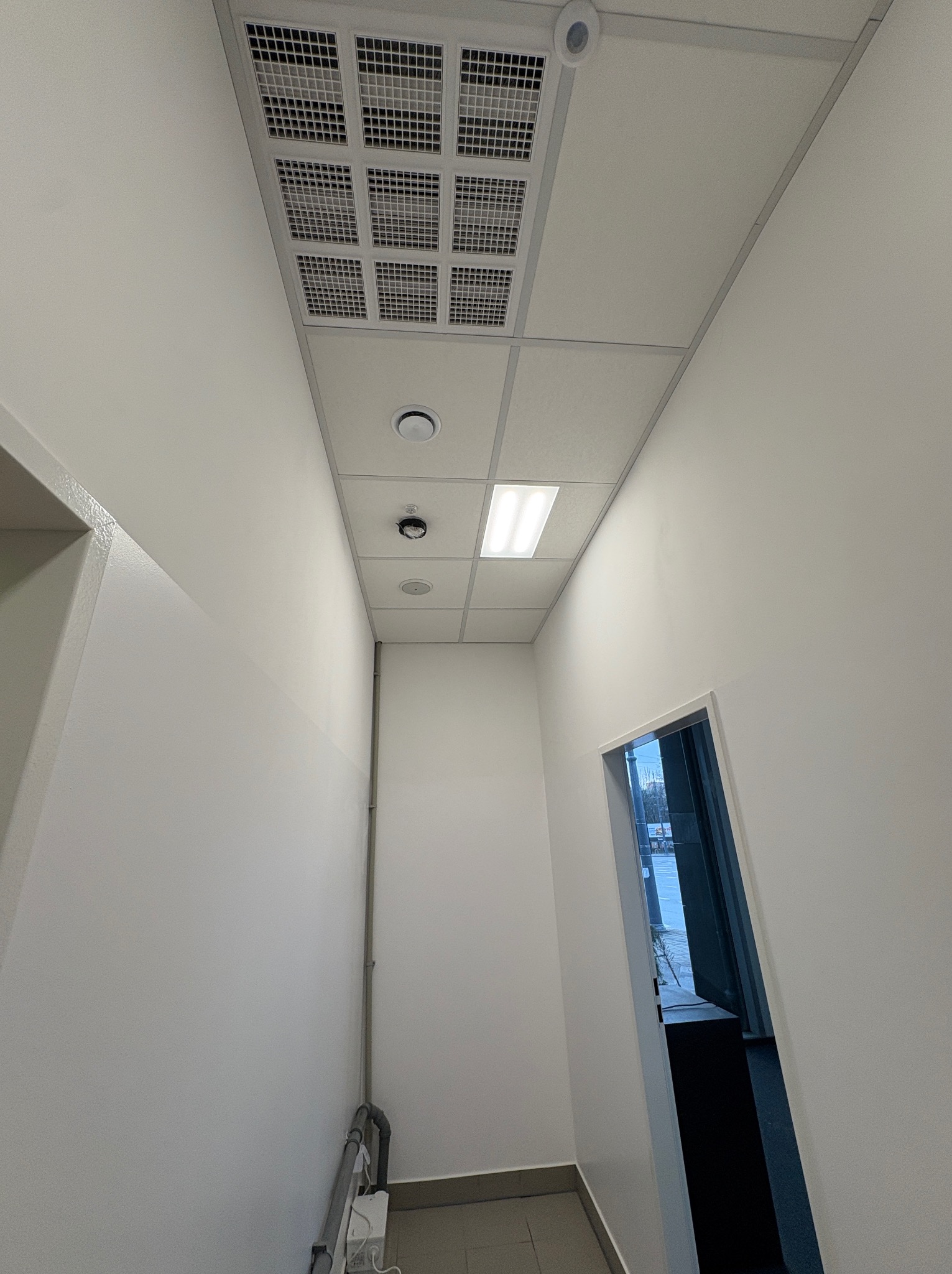 Białe wnętrze biurowe z kasetonowym sufitem podwieszanym, kratką wentylacyjną i oświetleniem LED. Widoczne świeżo pomalowane ściany i fragment instalacji.