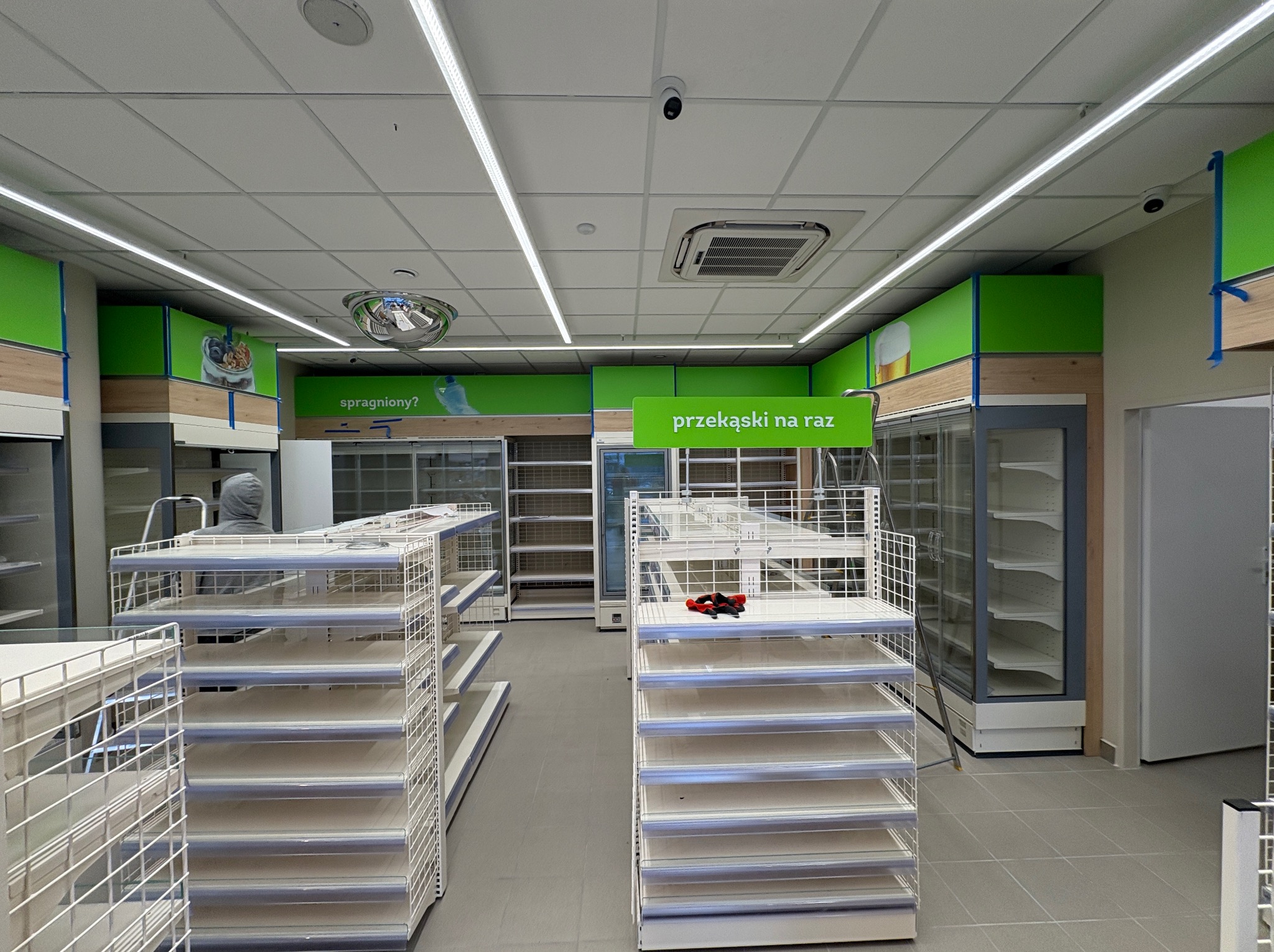Montaż wyposażenia sklepu: białe regały z siatki, chłodziarki i zielone panele dekoracyjne pod sufitem. Widoczne narzędzia i drabina.