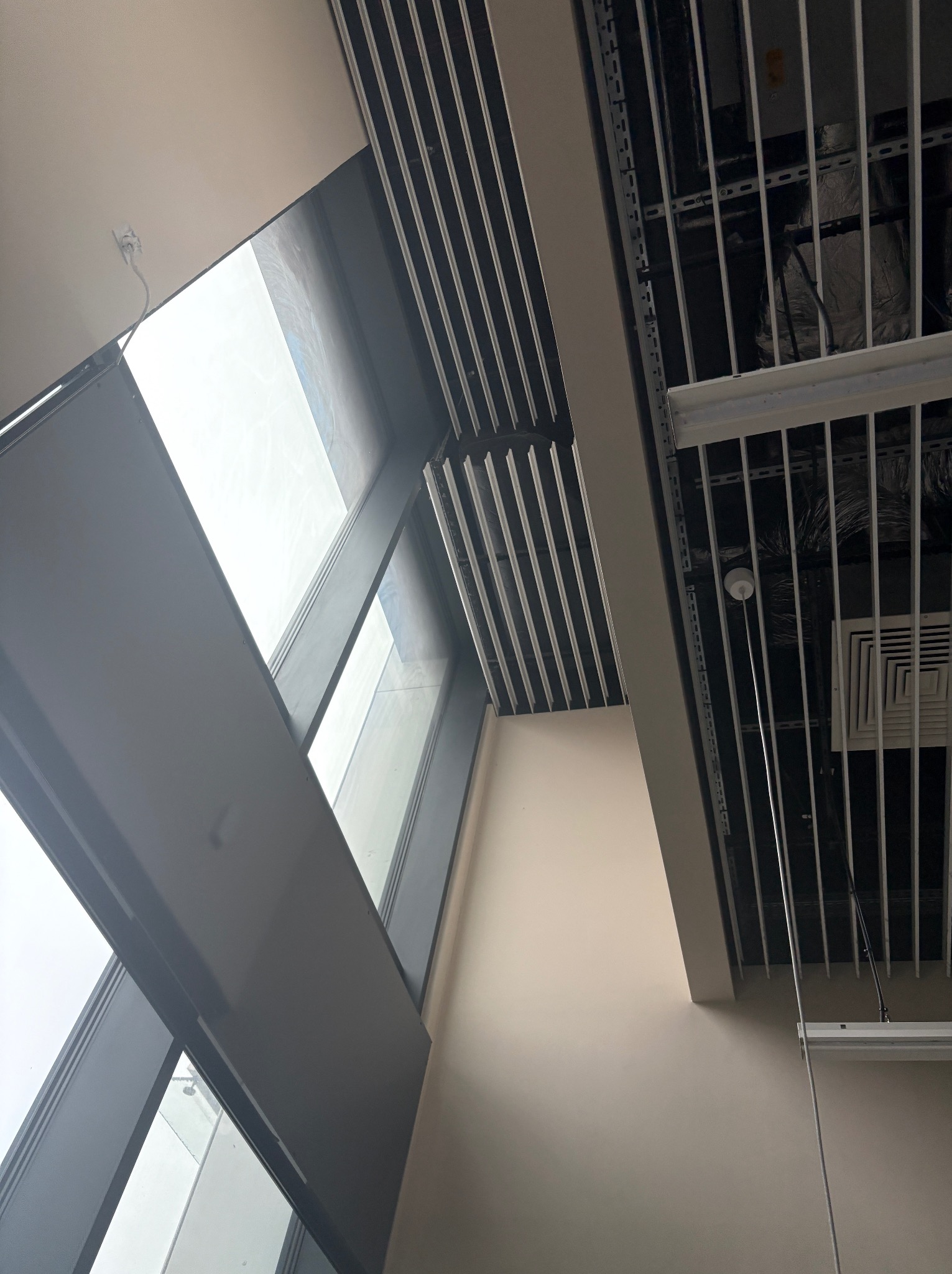 Nietypowy kadr na sufit biurowy z widoczną instalacją, oświetleniem i fragmentem okna. Widok z dołu, podkreślający geometrię i perspektywę wnętrza.