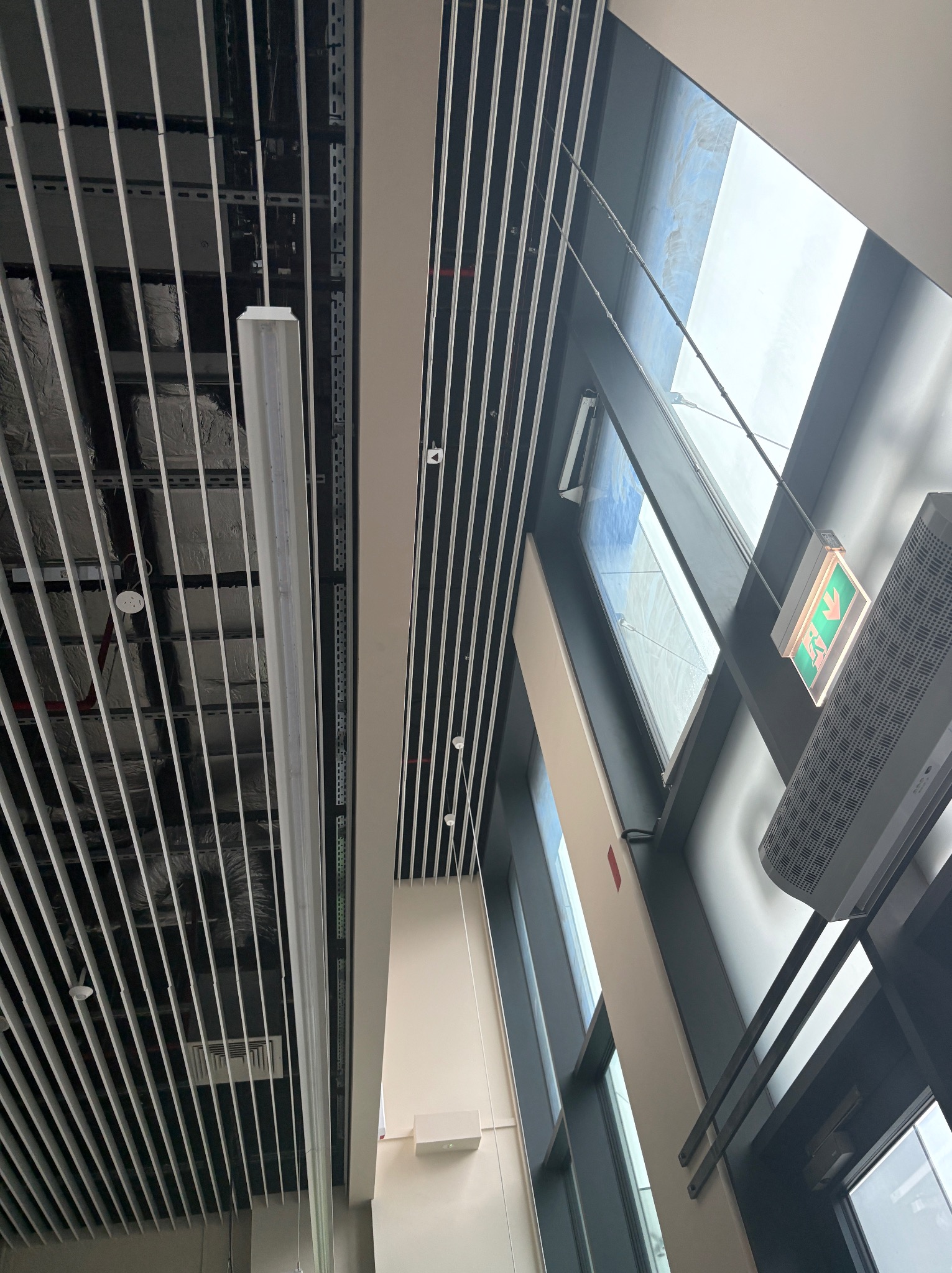 Nowoczesny sufit rastrowy z oświetleniem liniowym w biurze, widok z dołu, z fragmentem okna i klimatyzacji. Minimalistyczny design, białe elementy konstrukcyjne.