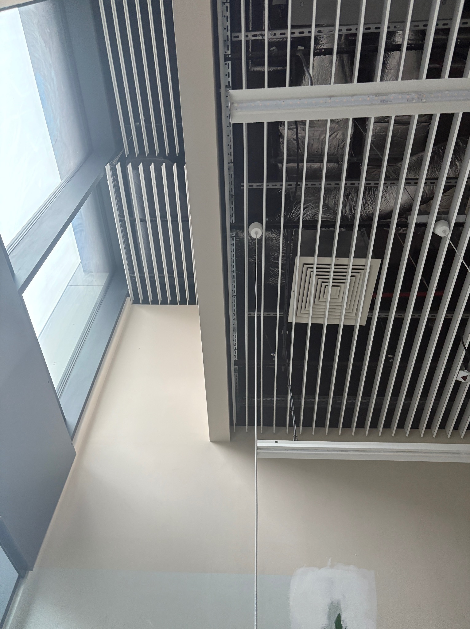 Biały sufit rastrowy z kratką wentylacyjną w biurze, widok z dołu, ściana pomalowana na jasny kolor, duże okno z widokiem na niebo. Oświetlenie LED.