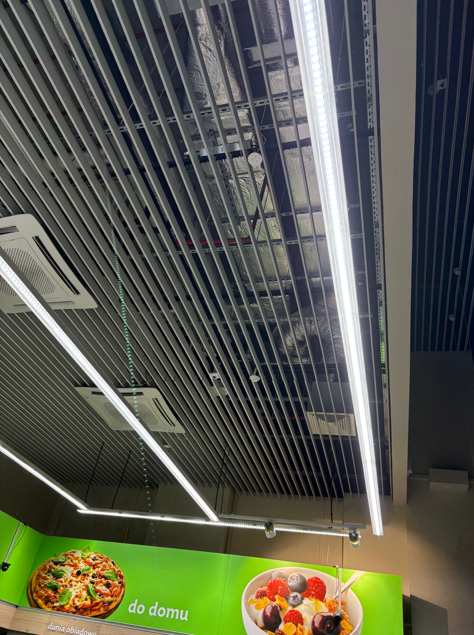 Sufit rastrowy z oświetleniem LED i klimatyzacją w przestrzeni komercyjnej. Widoczne elementy instalacji nad sufitem. Ujęcie z dołu, pod kątem.