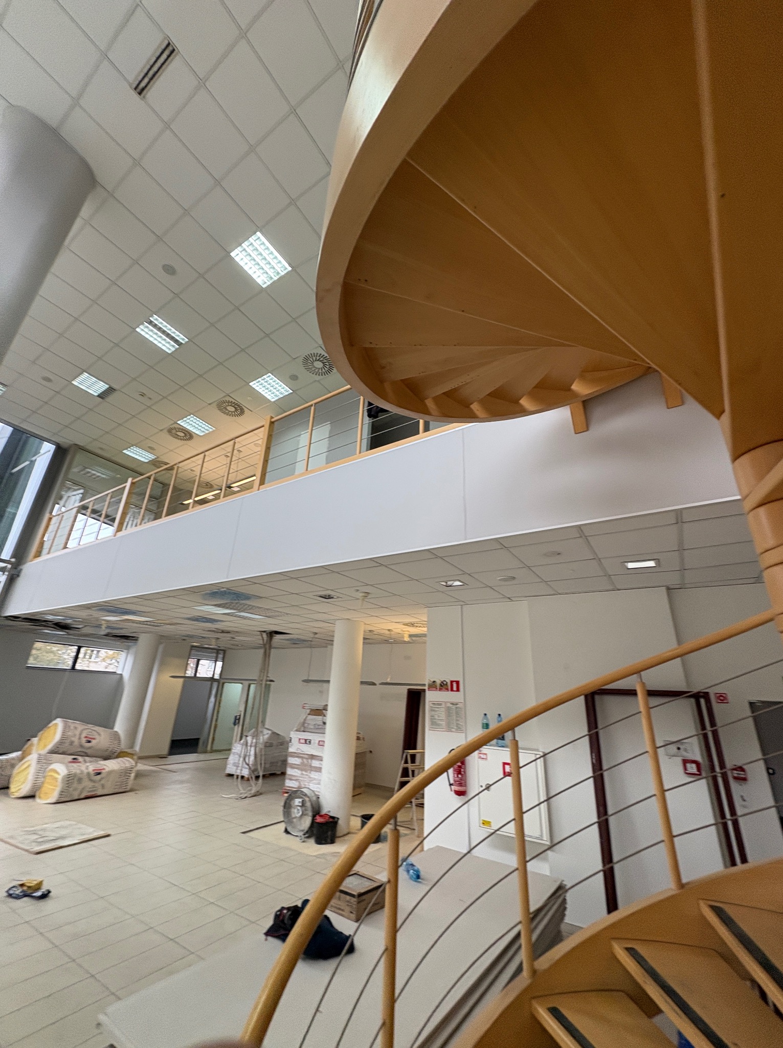 Wnętrze biura w trakcie prac wykończeniowych: widoczne materiały budowlane, drewniane schody spiralne i sufit kasetonowy. Ujęcie z dołu, podkreślające przestrzeń.