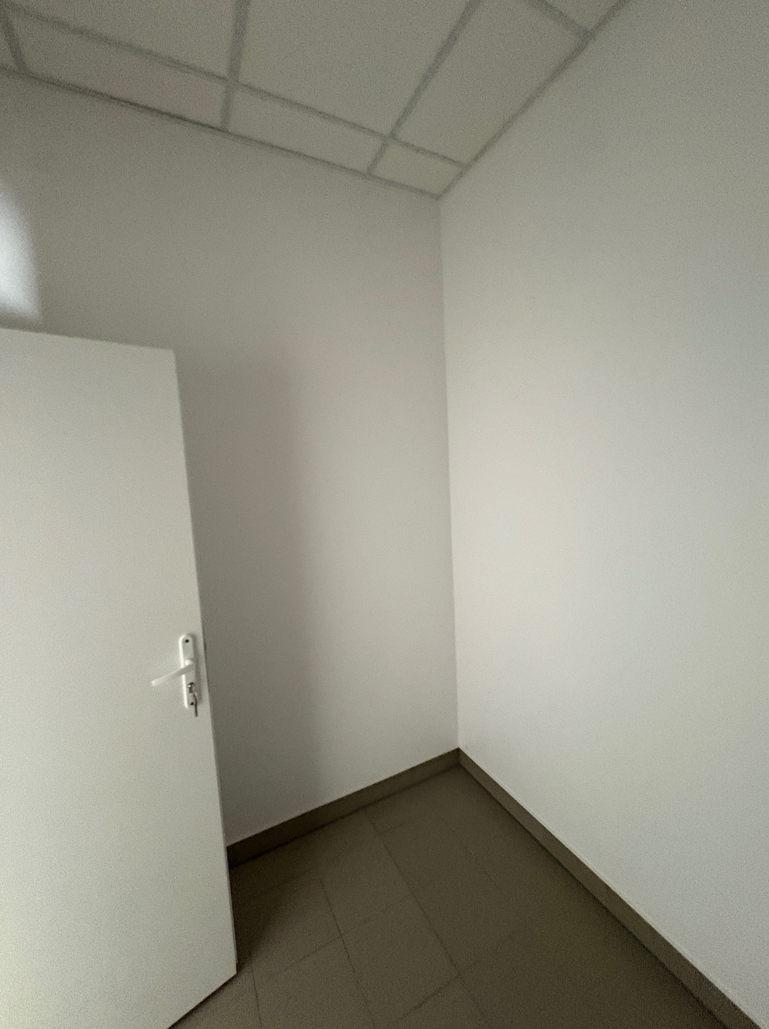 Pusty, świeżo pomalowany pokój biurowy z białą ścianą, drzwiami i szarą podłogą. Widoczny fragment sufitu kasetonowego. Minimalistyczne wnętrze po remoncie.