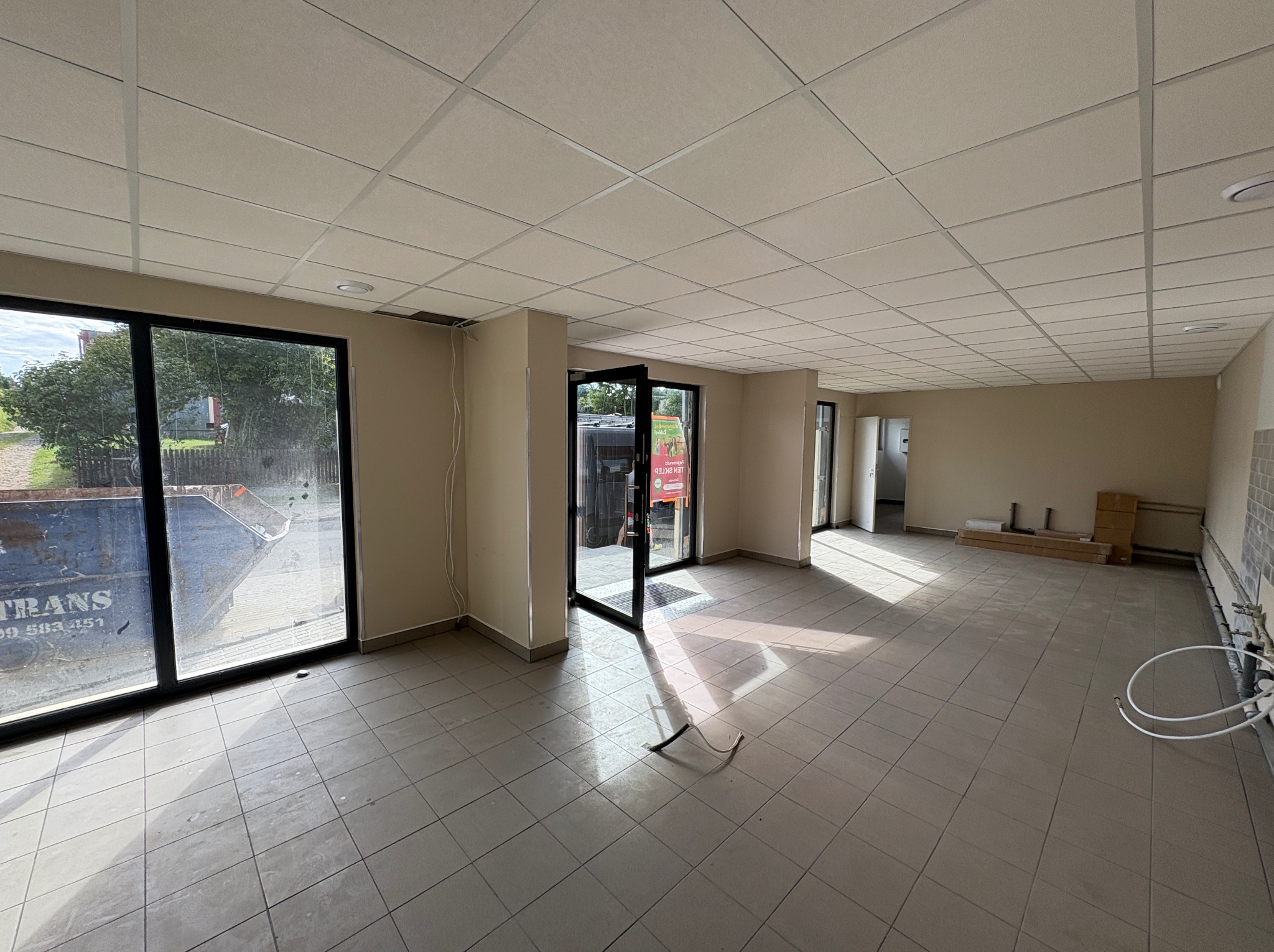 Wnętrze biura przed remontem: surowy stan z widoczną instalacją, płytkami podłogowymi i kasetonowym sufitem. Duże okna z widokiem na zewnątrz.