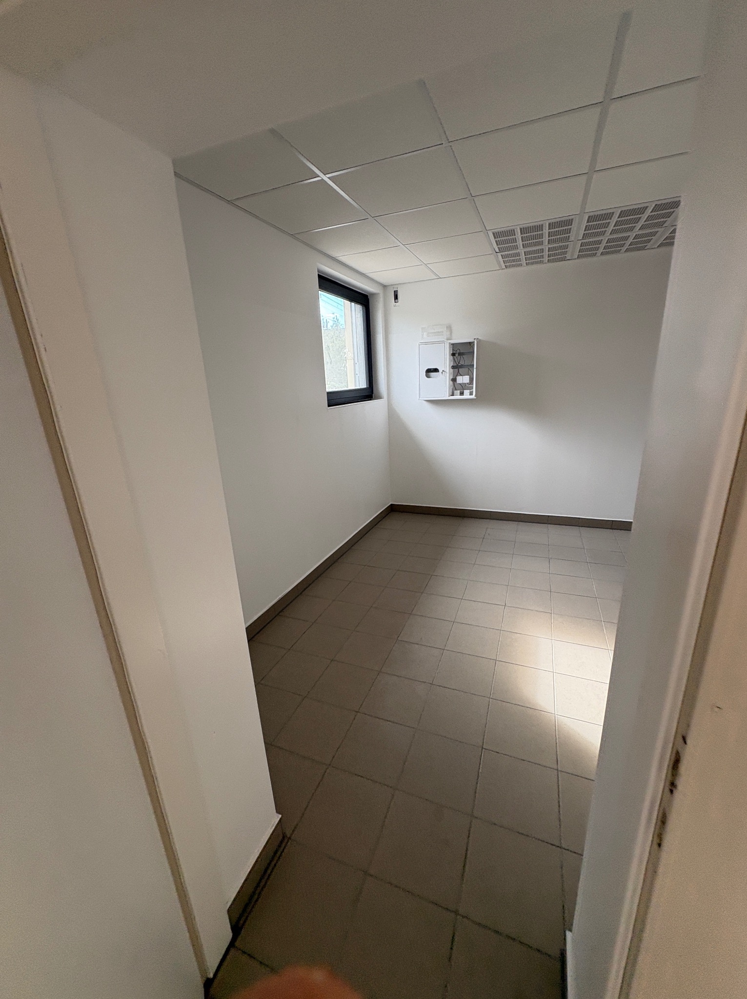 Wnętrze biura po remoncie: białe ściany, szara podłoga z płytek, okno z czarną ramą, sufit kasetonowy z kratką wentylacyjną i szafka elektryczna na ścianie.