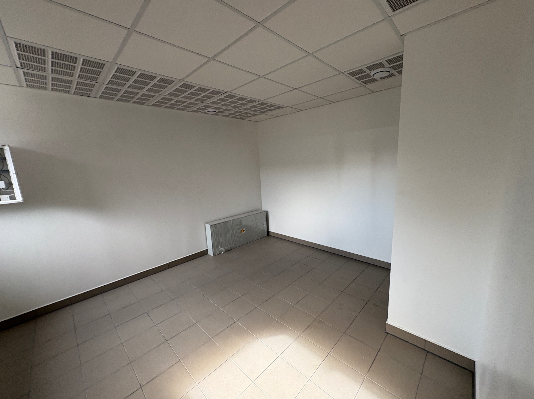 Puste biuro po remoncie: świeżo pomalowane ściany, płytki podłogowe, sufit kasetonowy z kratkami wentylacyjnymi. Grzejnik owinięty folią ochronną przy ścianie.