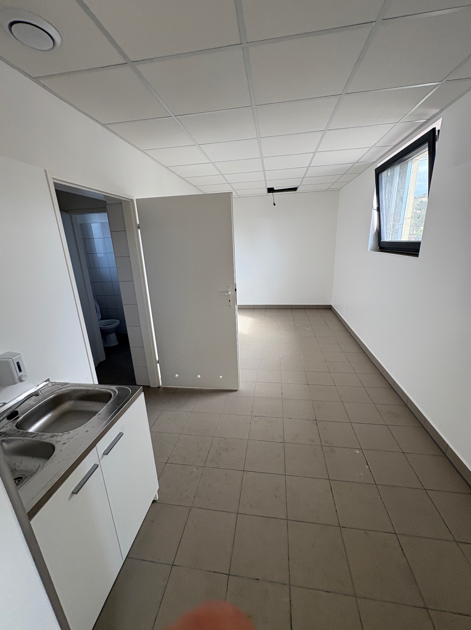 Wnętrze biurowe po remoncie: białe ściany, sufit kasetonowy, okno z żaluzjami, umywalka z szafką i widok na toaletę. Płytki podłogowe w neutralnym odcieniu.