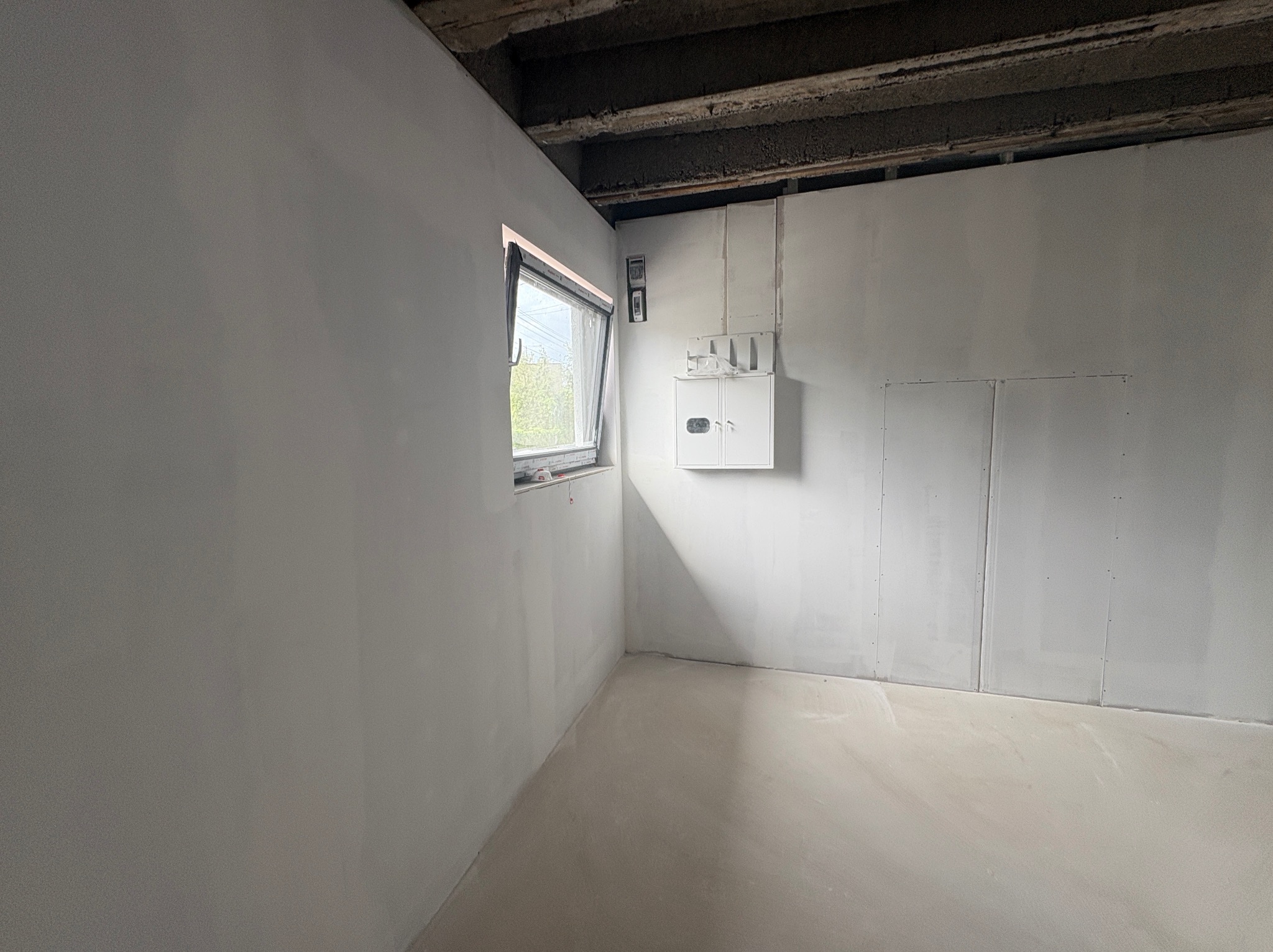 Wnętrze po szpachlowaniu ścian i wylewce podłogi, widoczne okno i skrzynka elektryczna. Surowy stan wykończenia, przygotowany do dalszych prac.