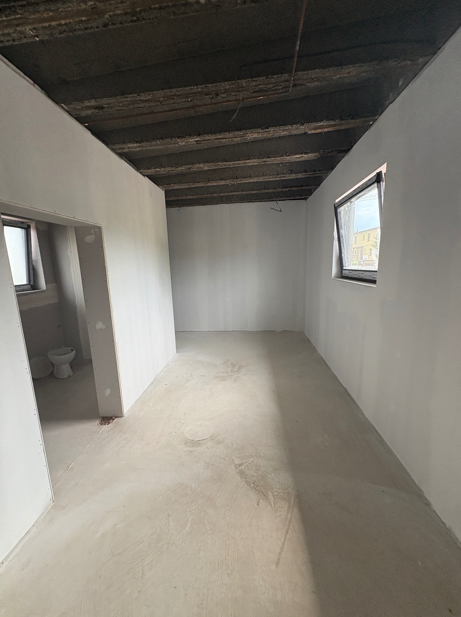 Wnętrze w trakcie prac wykończeniowych: surowy strop, ściany po szpachlowaniu, w tle widoczna toaleta w trakcie montażu. Okno z widokiem na budynek.