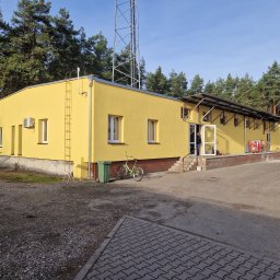 MichBud - Żółty budynek z anteną w tle, widok z zewnątrz. Na ścianie drabina, obok zaparkowany rower. Osoba w drzwiach. Teren wokół budynku utwardzony.