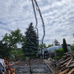 Wylewka fundamentów domu jednorodzinnego z użyciem pompy do betonu. Widoczne zbrojenie, szalunki i miksokret. Prace budowlane w toku, drzewa w tle.