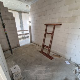 Wnętrze w trakcie budowy: ściany z białych bloczków, metalowe podpory, rozłożone rusztowanie. Surowy betonowy sufit z widocznymi instalacjami. Etap wznoszenia ścian działowych.