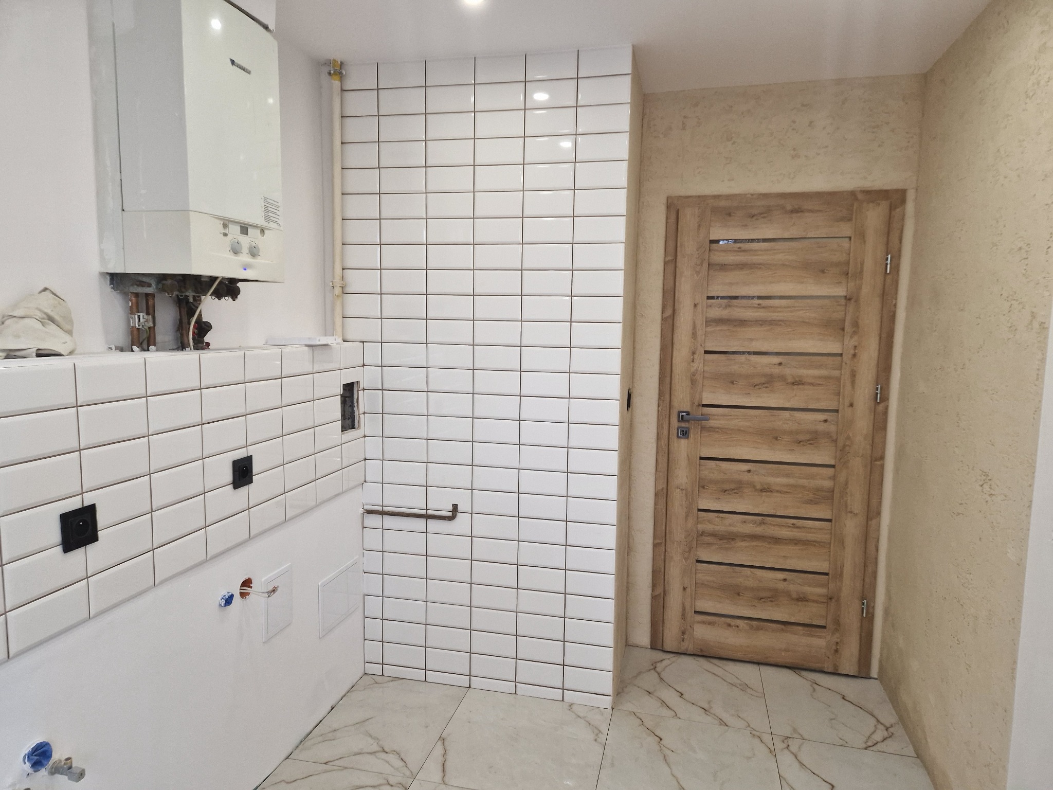 Wykończona łazienka z białymi, prostokątnymi płytkami, drewnianymi drzwiami i marmurową podłogą. Widoczny piec gazowy i instalacje. Ściana pokryta dekoracyjnym tynkiem.