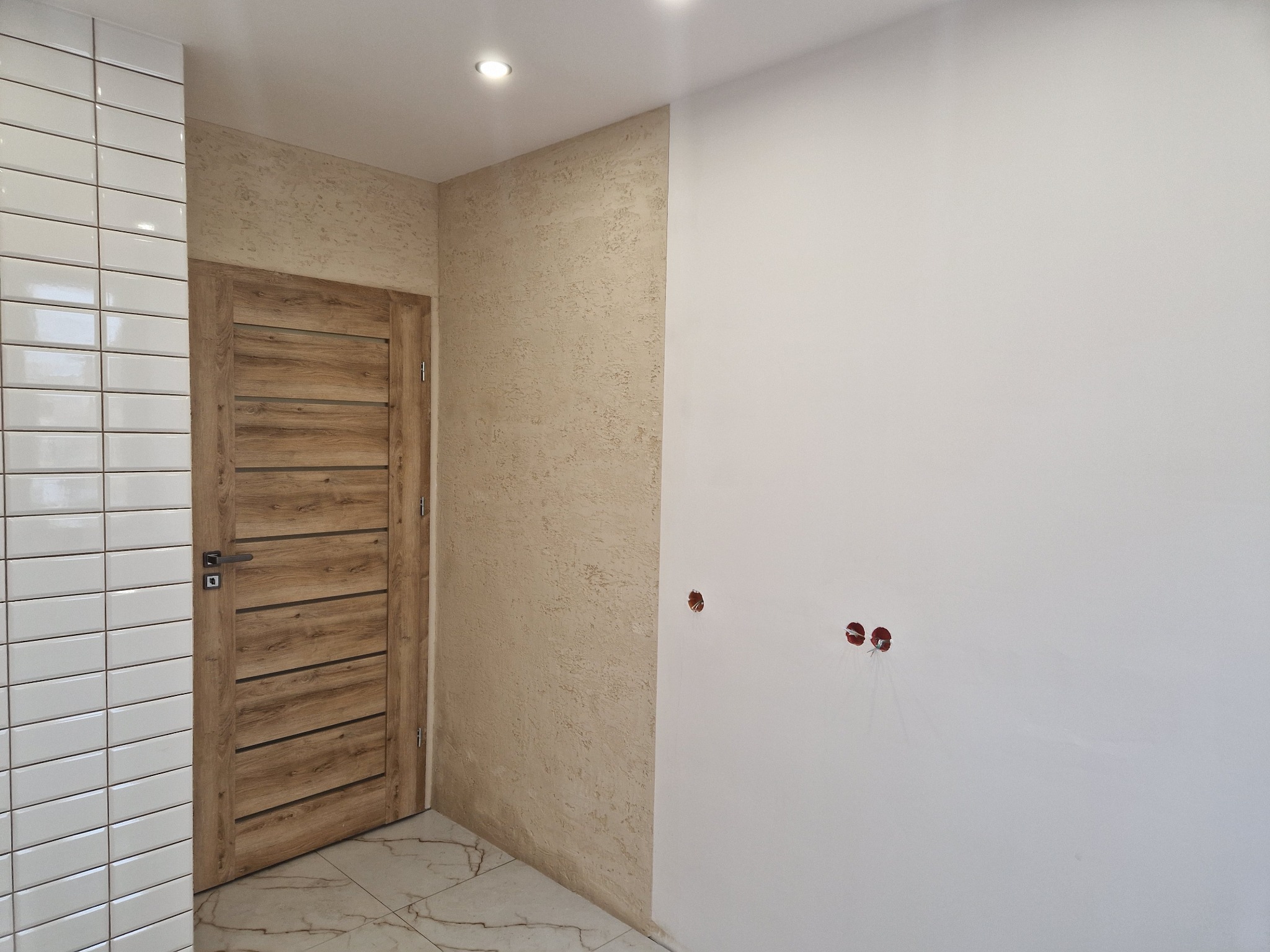 Wnętrze po remoncie: drewniane drzwi z poziomymi panelami, białe płytki na ścianie, ściana z beżową fakturą i biała ściana z puszkami elektrycznymi. Marmurowa podłoga.