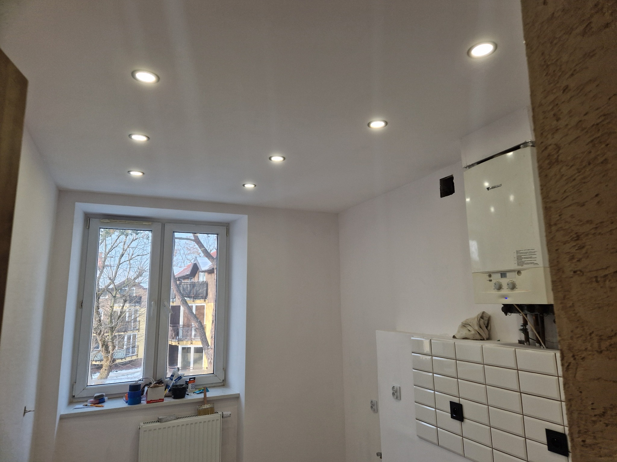 Białe wnętrze po remoncie: świeżo pomalowane ściany, sufit z halogenami, okno z widokiem na budynek, grzejnik i fragment kotła gazowego z białą glazurą.