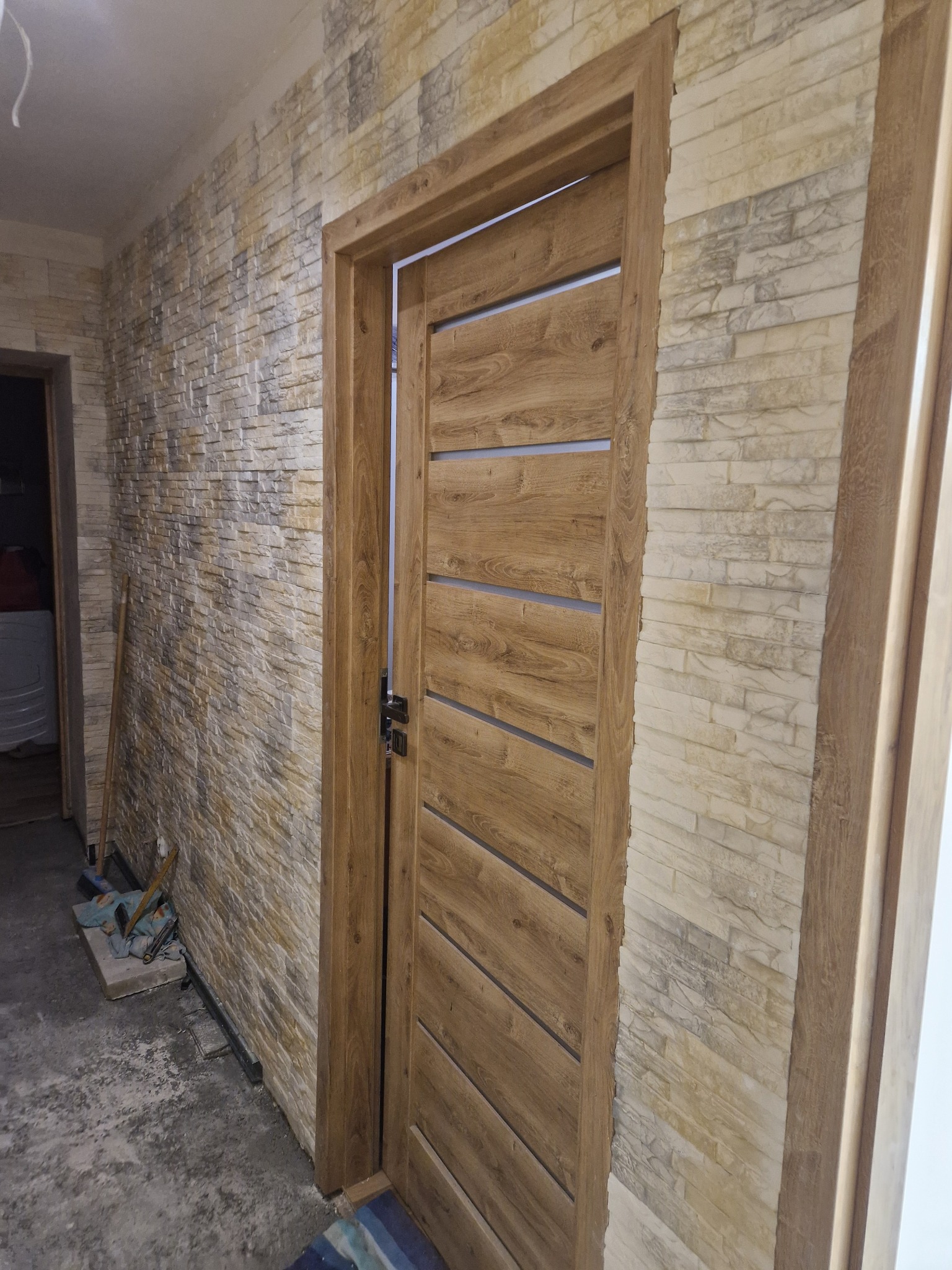 Zamontowane drewniane drzwi wewnętrzne z poziomymi przeszkleniami, dopasowane do ściany wykończonej kamieniem dekoracyjnym w odcieniach beżu i szarości. Widoczne narzędzia na podłodze.
