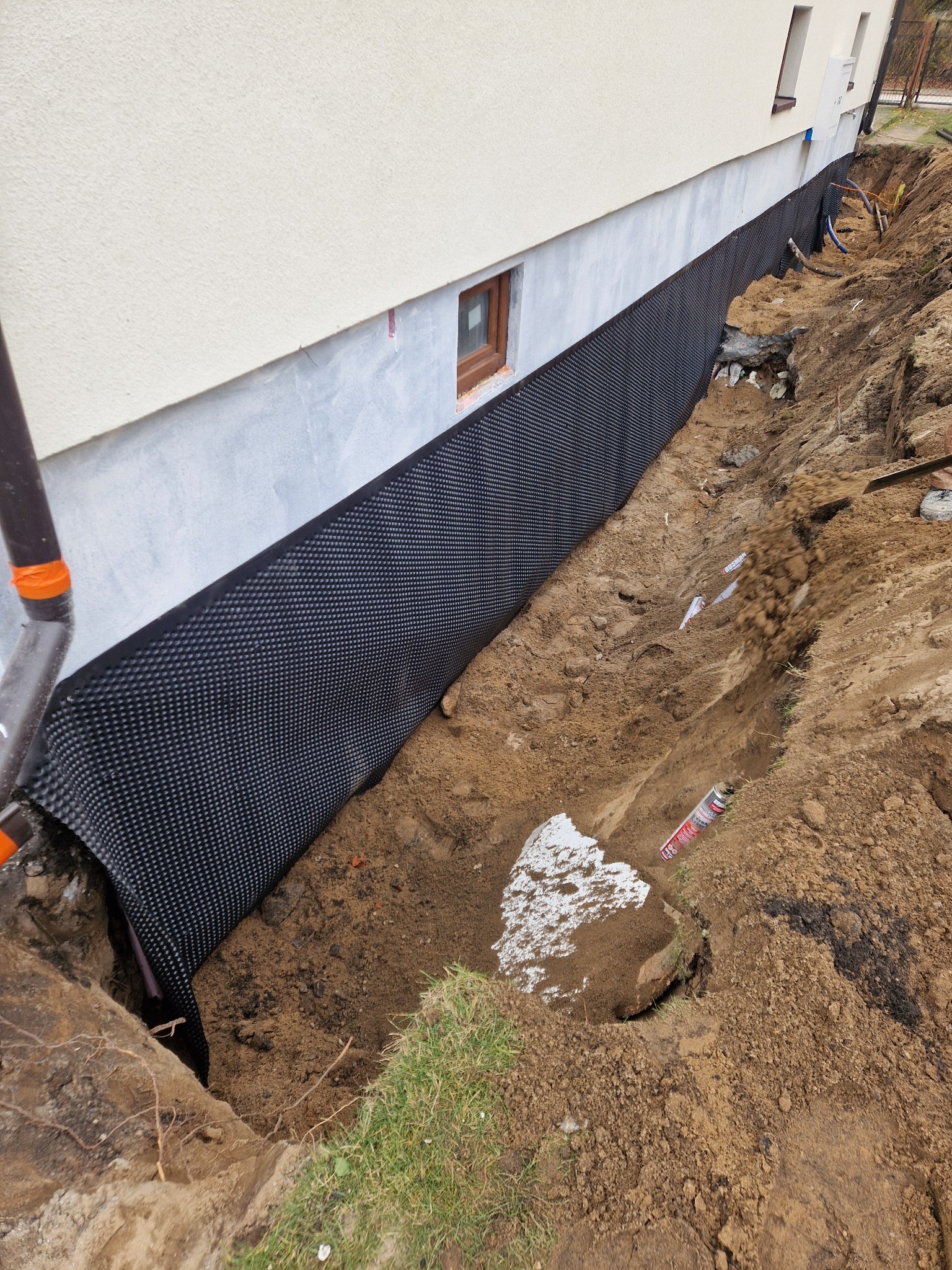 Fundament budynku zabezpieczony czarną folią kubełkową w wykopie. Widoczne okno piwniczne i fragment elewacji. Prace ziemne w toku, częściowo zasypane.