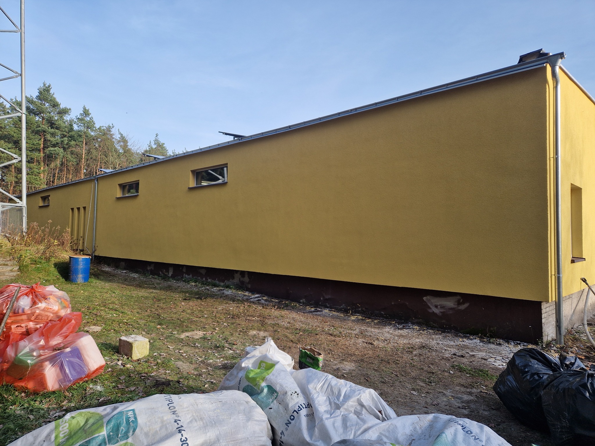 Ocieplony budynek w odcieniach żółci z widocznymi workami materiałów budowlanych i niebieską beczką, na tle drzew i błękitnego nieba.