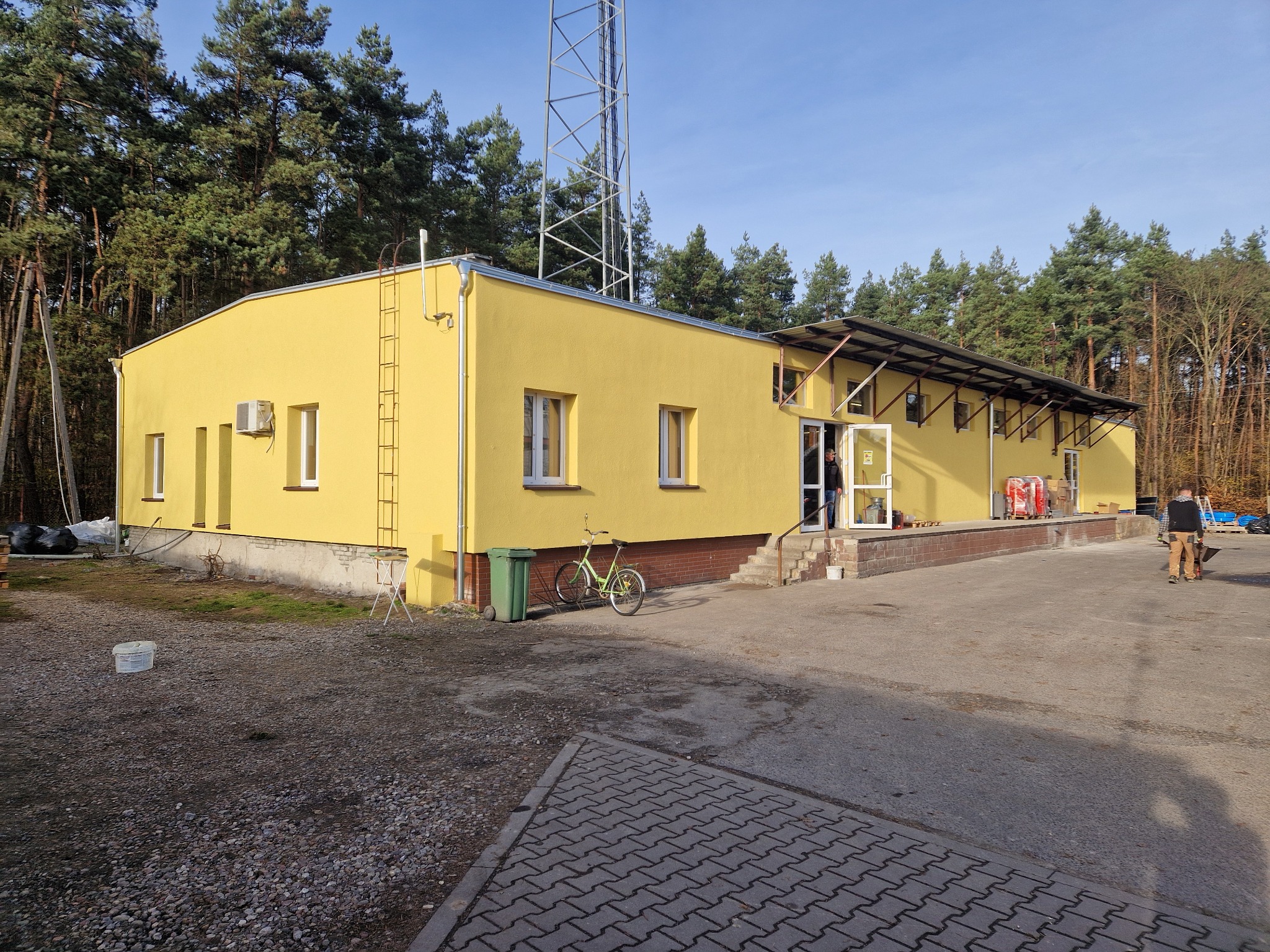 Żółty budynek z anteną w tle, widok z zewnątrz. Na ścianie drabina, obok zaparkowany rower. Osoba w drzwiach. Teren wokół budynku utwardzony.