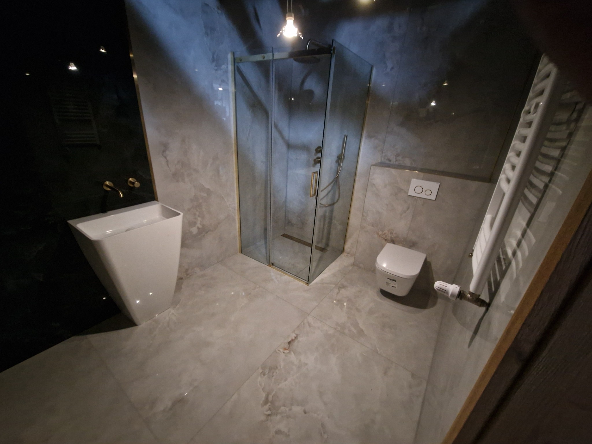 Nowoczesna łazienka z kabiną prysznicową, umywalką na postumencie i grzejnikiem drabinkowym. Szare płytki imitujące marmur, złote akcenty. Oświetlenie punktowe.