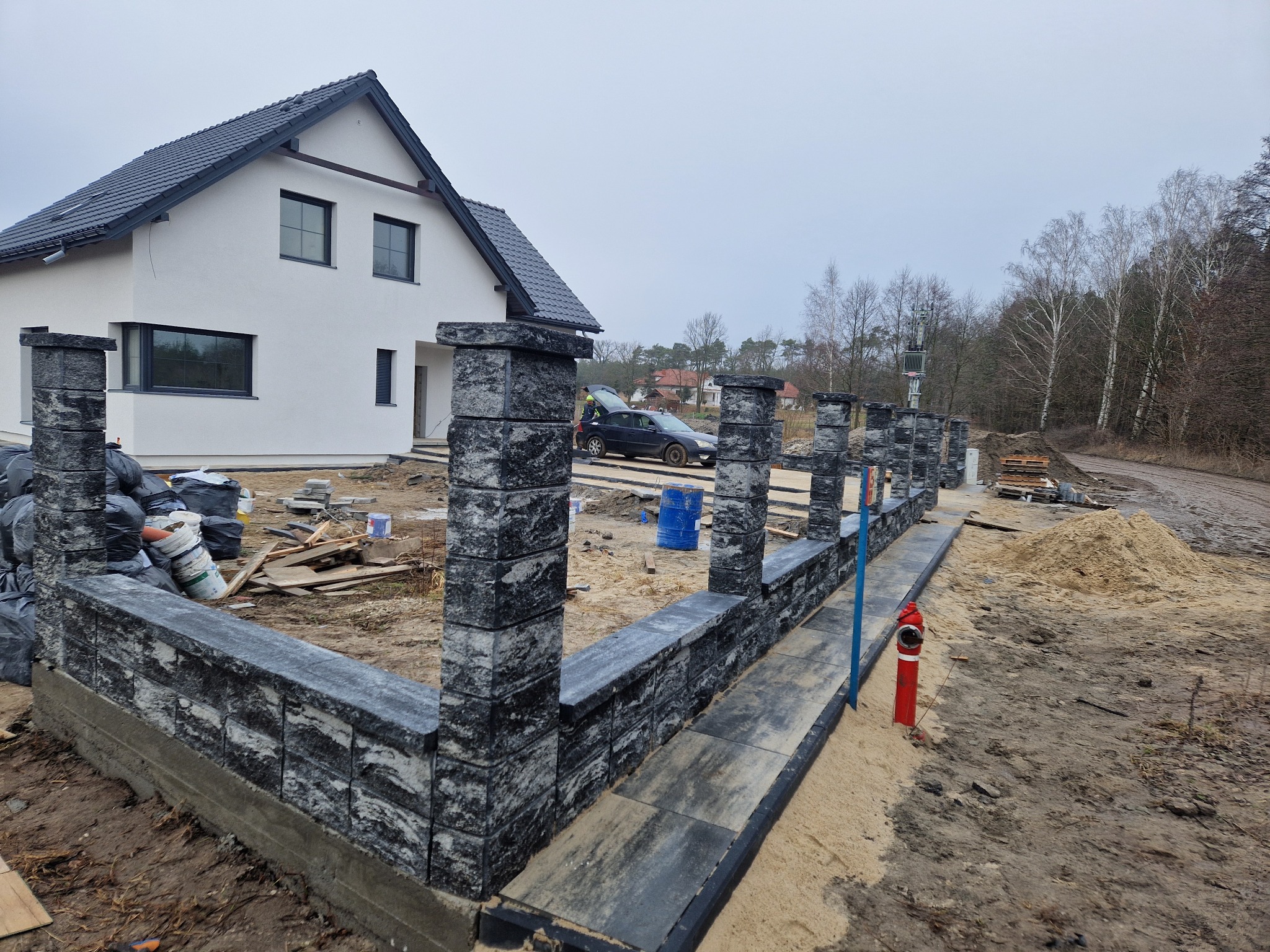 Wznoszony mur z szarej kostki granitowej, widoczny na tle białego domu z ciemnym dachem i drzew. Na pierwszym planie czerwony hydrant i piasek.