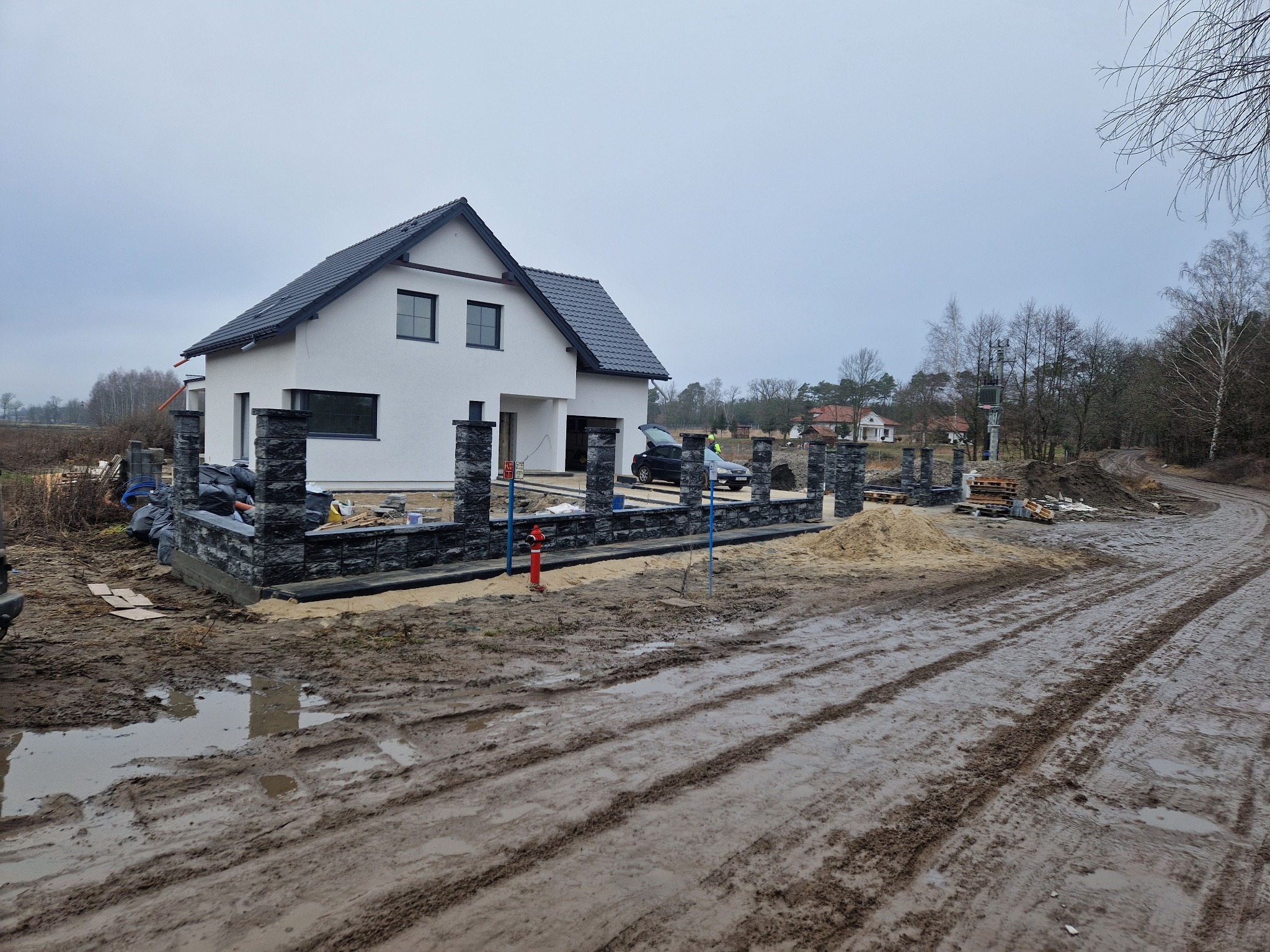 Nowoczesny, biały dom z ciemnym dachem i ogrodzeniem w trakcie budowy. Widoczna błotnista droga dojazdowa i materiały budowlane. Pochmurne niebo.