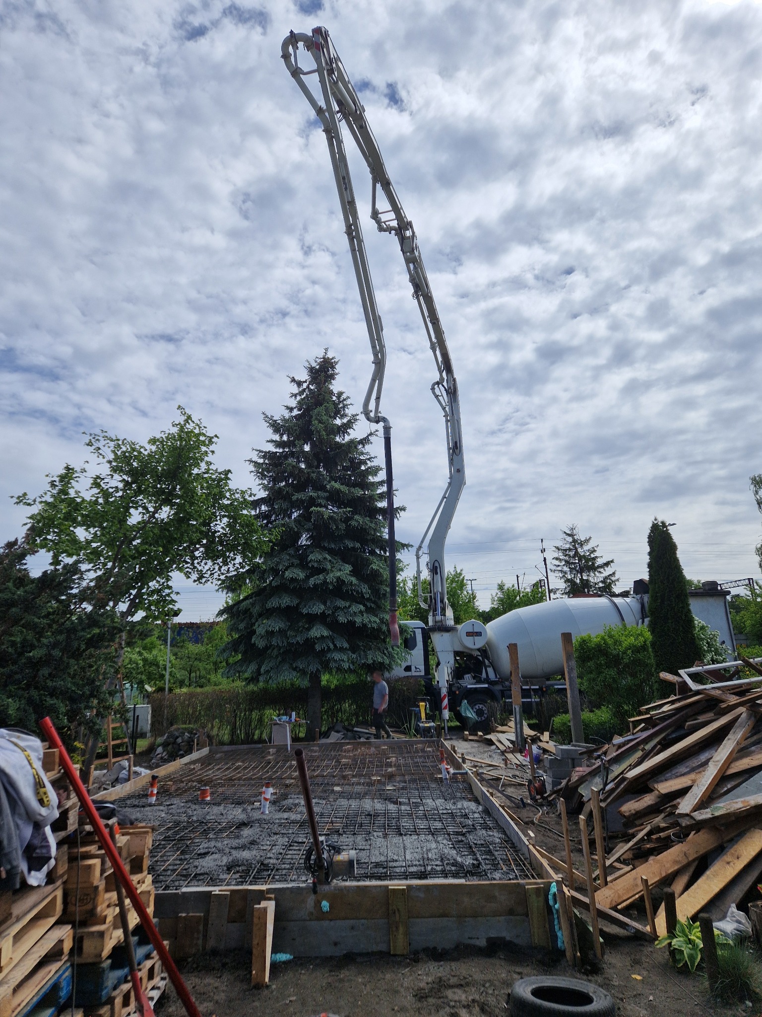 Wylewka fundamentów domu jednorodzinnego z użyciem pompy do betonu. Widoczne zbrojenie, szalunki i miksokret. Prace budowlane w toku, drzewa w tle.