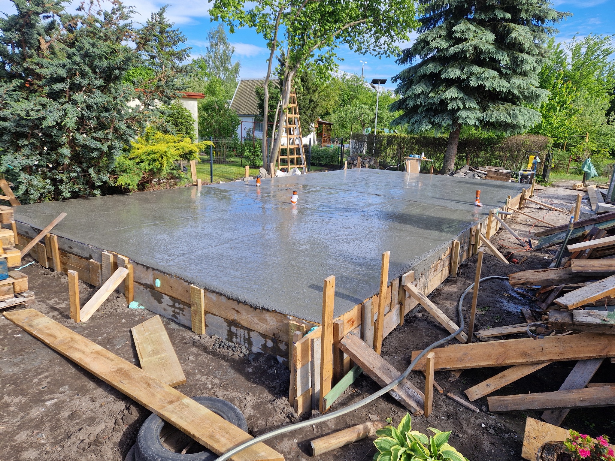 Wylany fundament pod budowę domu jednorodzinnego, widoczne szalunki i wystające rury instalacyjne. W tle zieleń drzew i częściowo widoczny budynek.