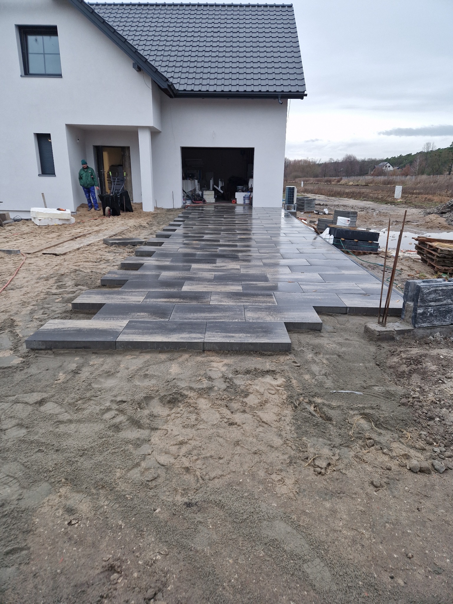 Układanie szarej kostki brukowej przed nowoczesnym domem z białymi ścianami i grafitowym dachem. Widoczny fragment posesji w trakcie prac.