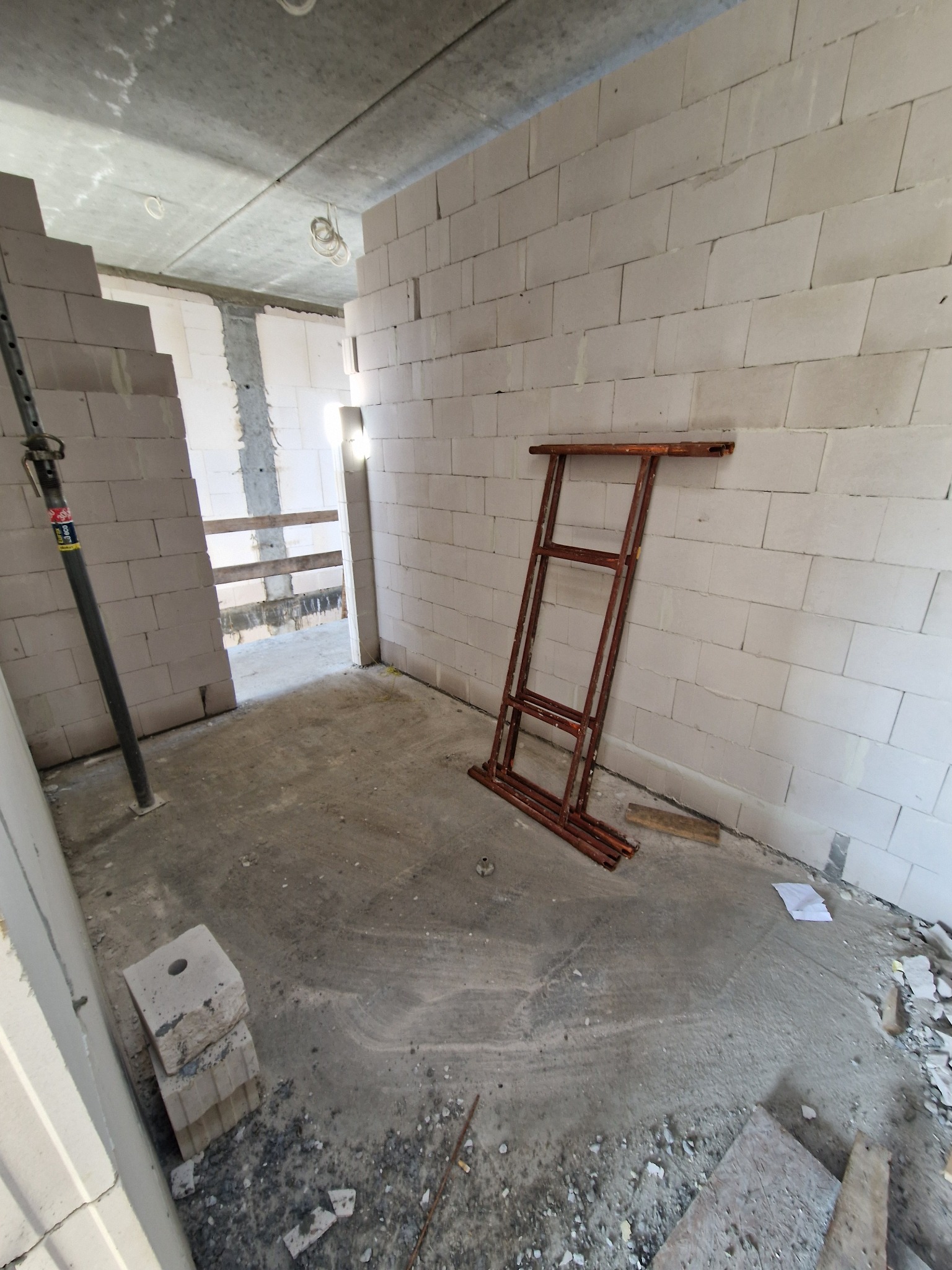 Wnętrze w trakcie budowy: ściany z białych bloczków, metalowe podpory, rozłożone rusztowanie. Surowy betonowy sufit z widocznymi instalacjami. Etap wznoszenia ścian działowych.