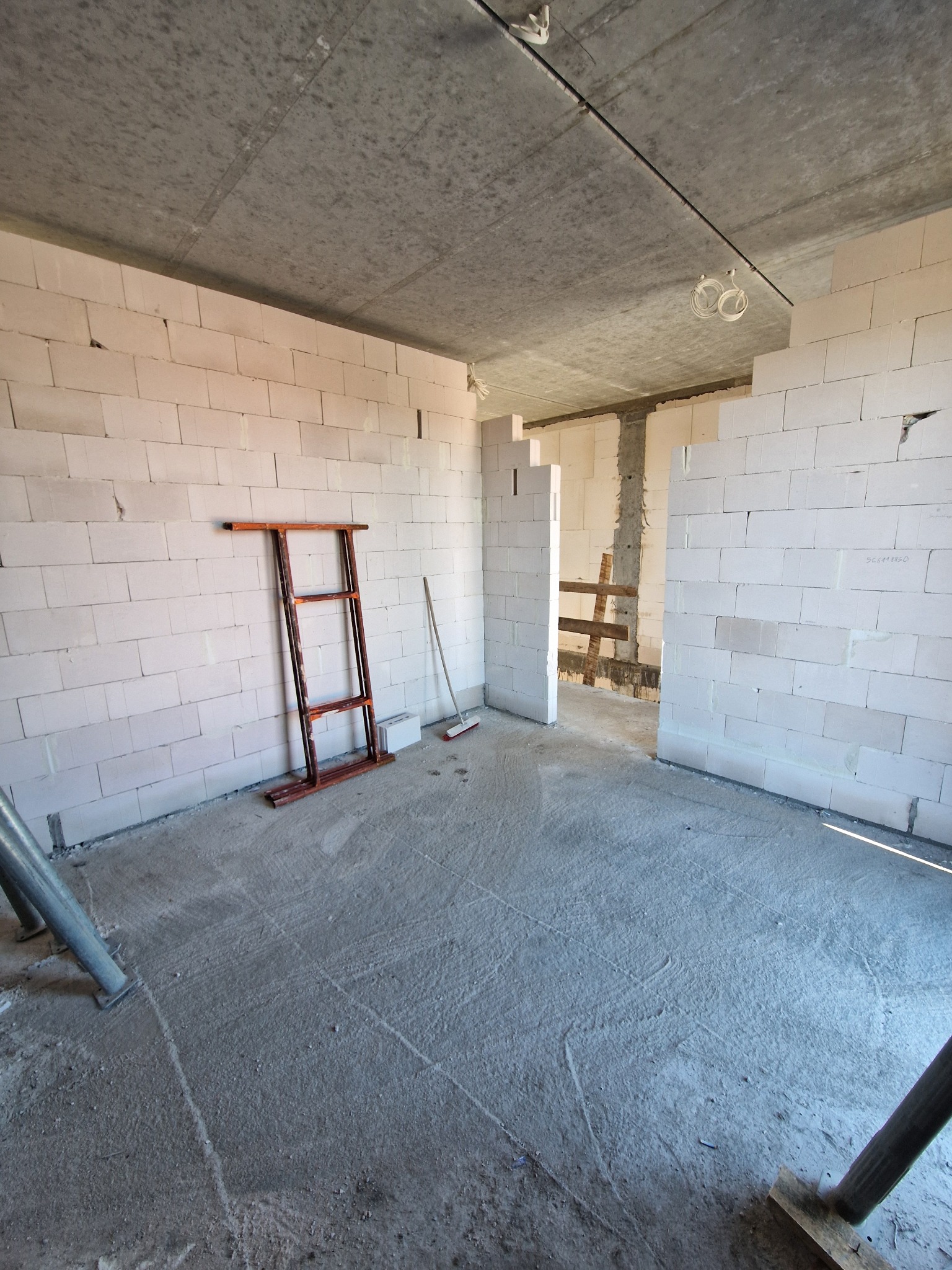Surowy stan wnętrza: ściany z białych bloczków, surowy betonowy sufit, metalowe podpory i drabina. W tle widoczne kolejne pomieszczenie w trakcie budowy.