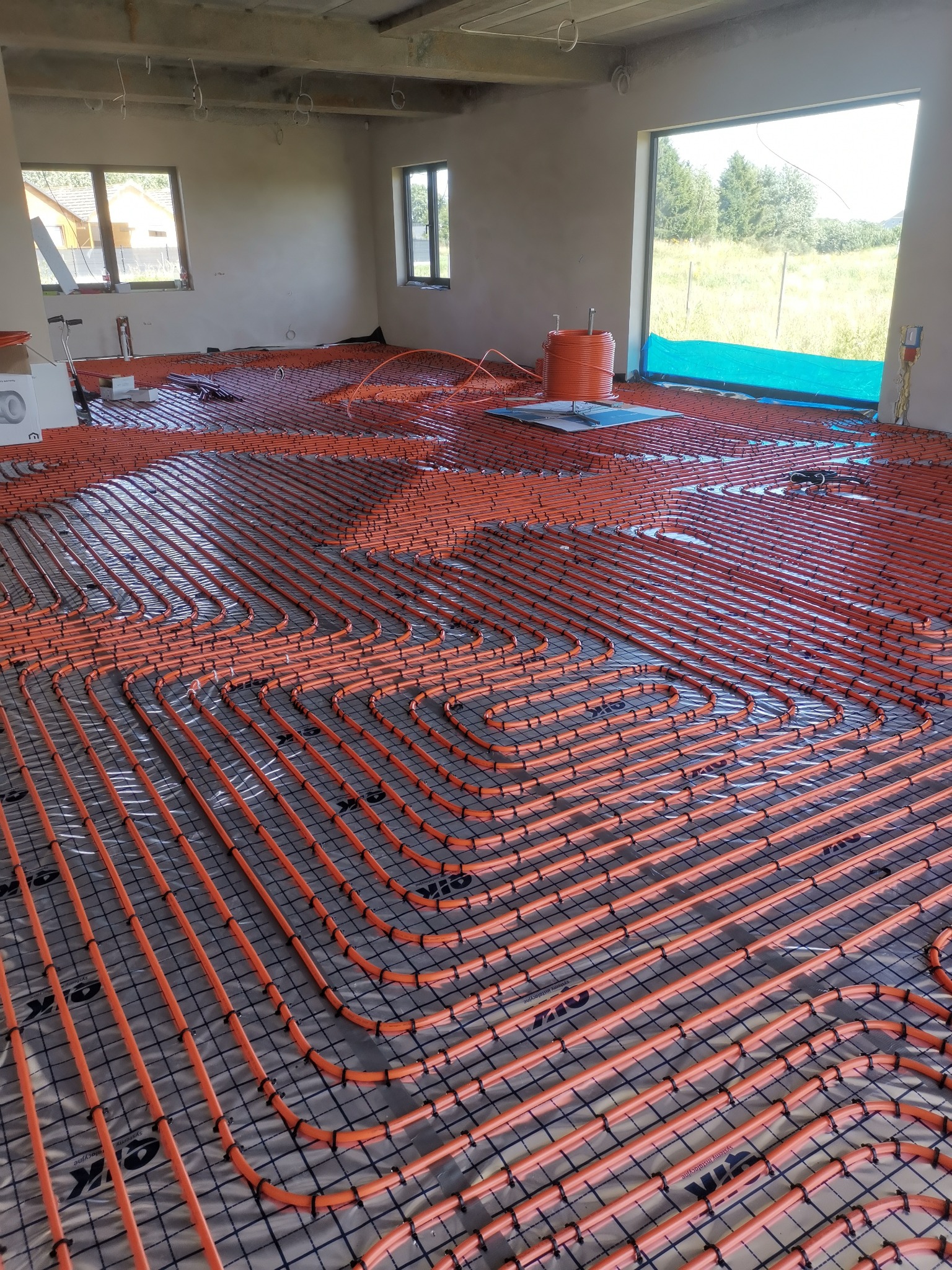 Pomarańczowe rury ogrzewania podłogowego ułożone w pętle na folii izolacyjnej w surowym wnętrzu z oknami. Widoczny rozdzielacz i elementy montażowe.