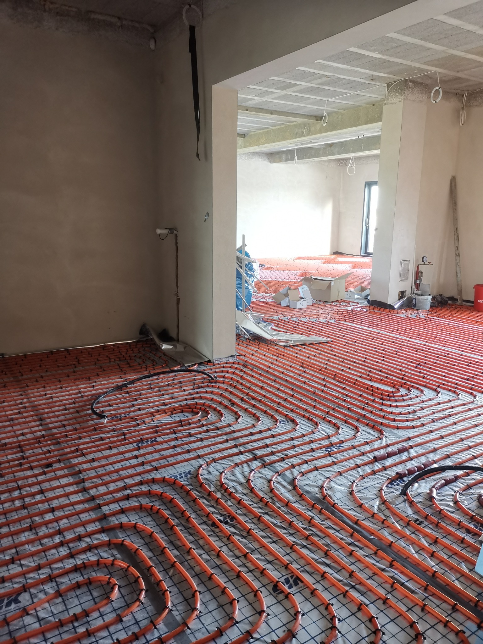Pomarańczowe rury ogrzewania podłogowego ułożone na folii z siatką w surowym wnętrzu. Widoczne materiały budowlane i instalacje pod sufitem.