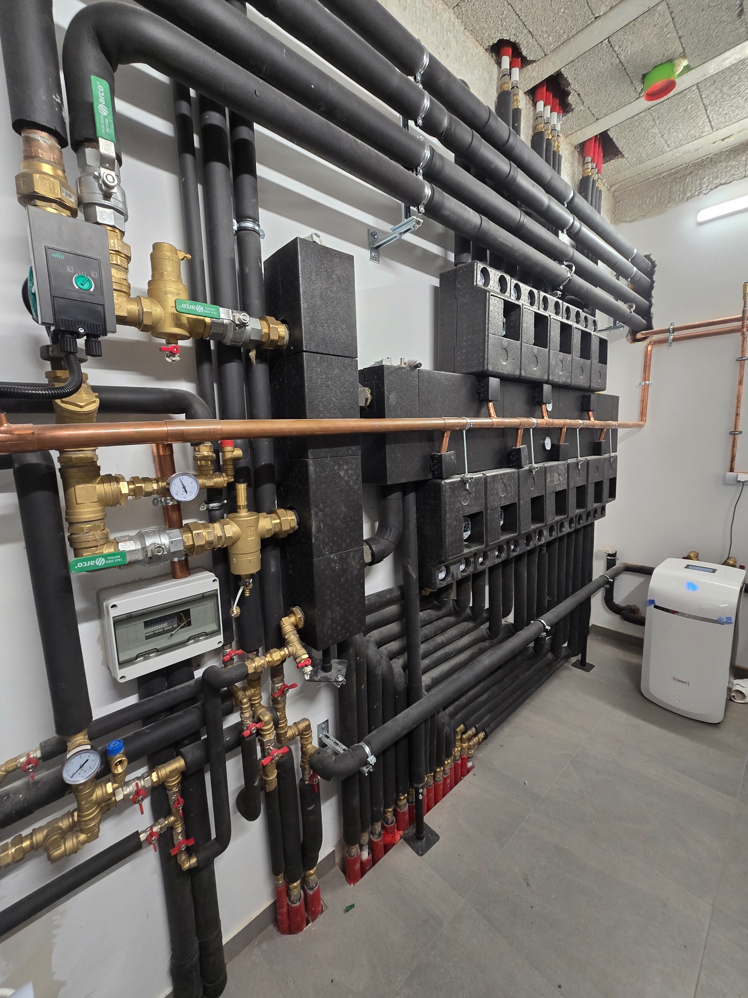 Złożona instalacja grzewcza z czarnymi rurami izolowanymi, mosiężnymi zaworami i rozdzielaczami, zamontowana na białej ścianie w pomieszczeniu technicznym.