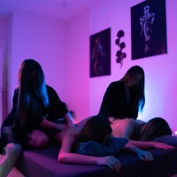 Cosmos Massage - Dwie osoby wykonują masaż pleców klientki na łóżku w pokoju oświetlonym fioletowym światłem. Na ścianie wiszą obrazy.