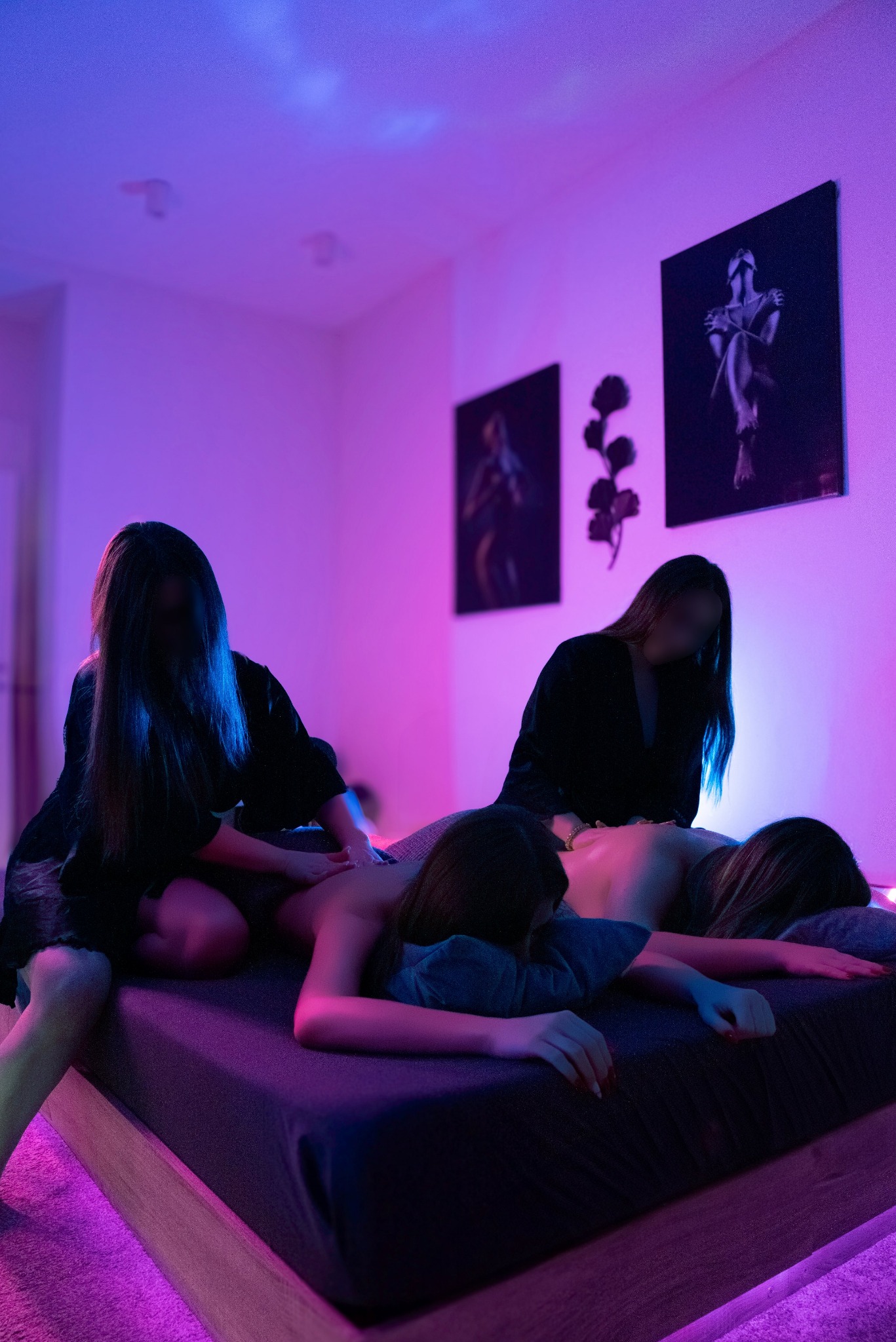 Dwie osoby wykonują masaż pleców klientki na łóżku w pokoju oświetlonym fioletowym światłem. Na ścianie wiszą obrazy.