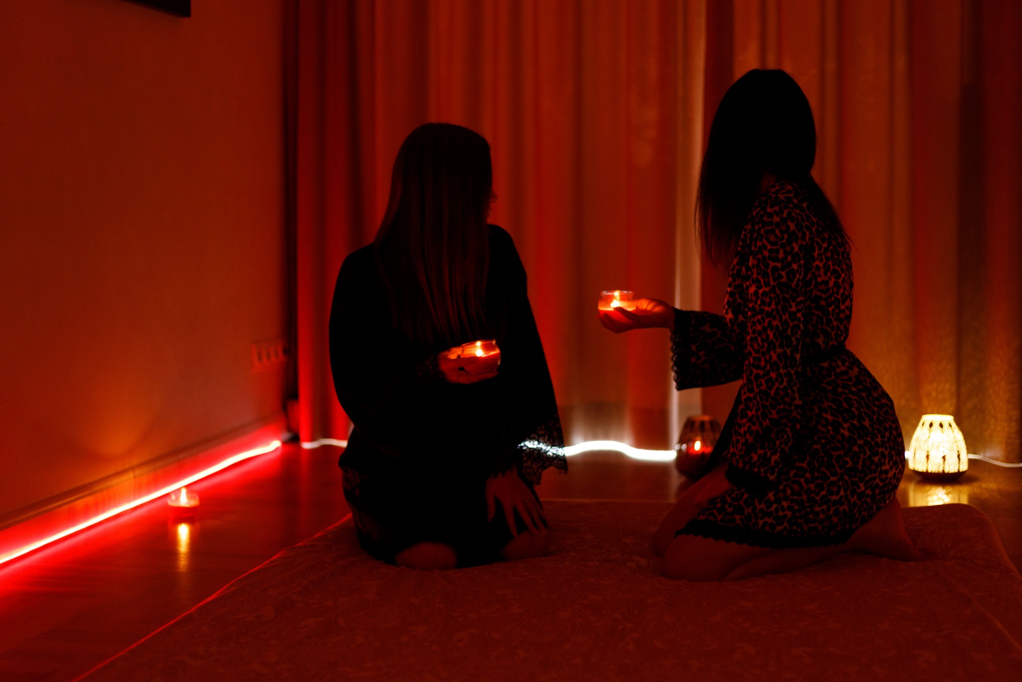 Dwie kobiety klęczą na macie w pokoju oświetlonym czerwonym światłem, trzymając zapalone świece w dłoniach. Klimatyczne oświetlenie i relaksująca atmosfera.