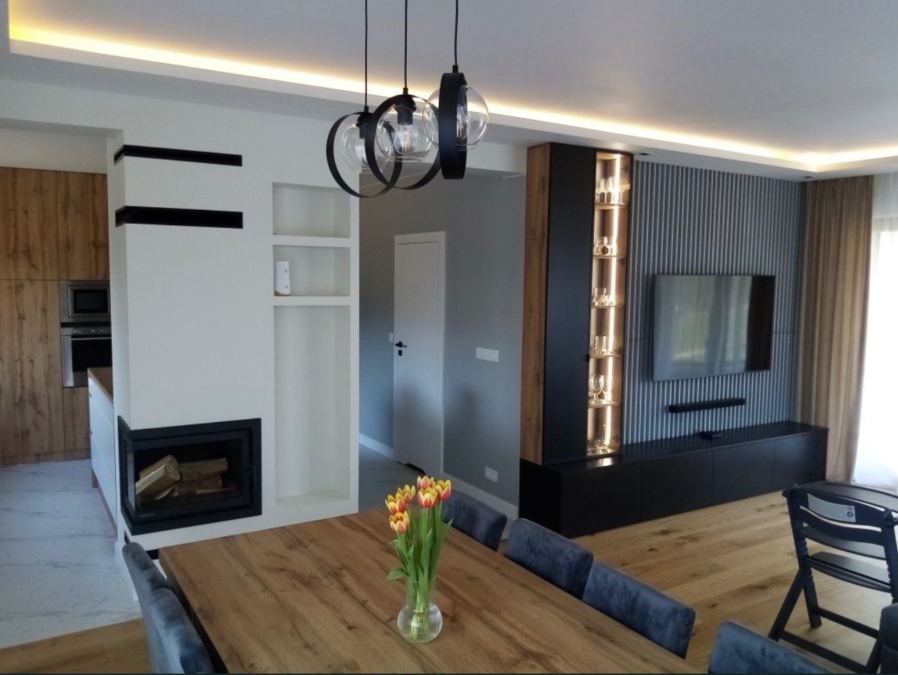 Nowoczesne wnętrze: drewniany stół z tulipanami, kominek, zabudowa TV z oświetleniem, drewniana podłoga i designerskie lampy wiszące. Minimalistyczny design.