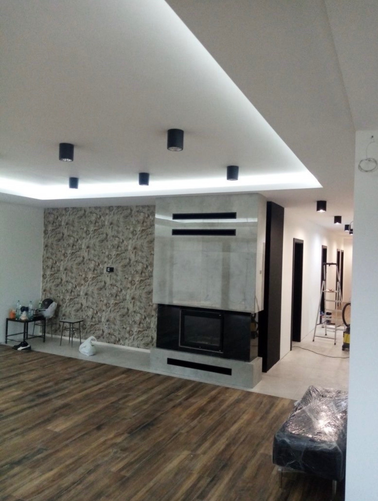 Nowoczesny salon po remoncie: drewniana podłoga, ściana z tapetą, kominek obudowany szarym kamieniem, oświetlenie sufitowe punktowe i ledowe. W tle drabina malarska.