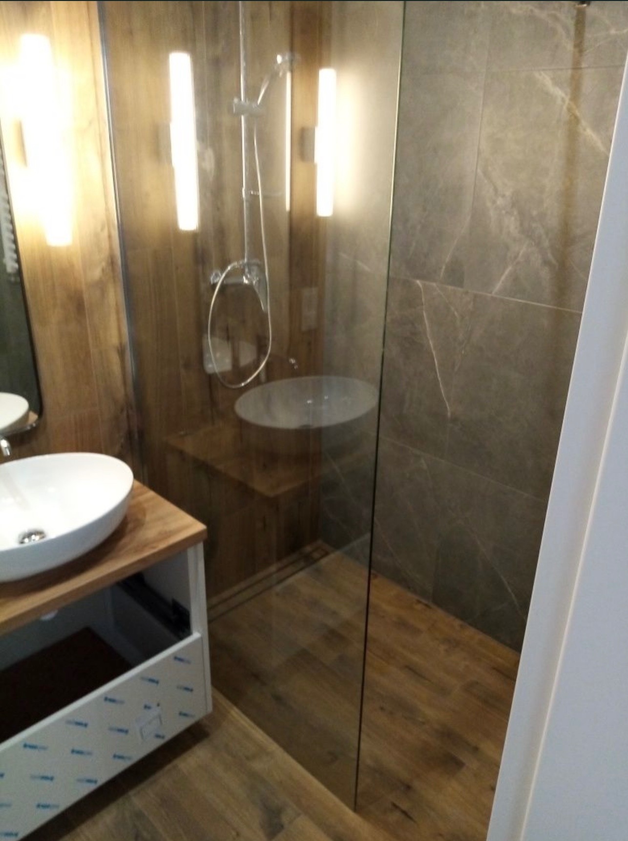 Nowoczesna łazienka z kabiną prysznicową typu walk-in, umywalką nablatową na drewnianym blacie i oświetleniem LED. Widoczne płytki imitujące drewno i marmur.